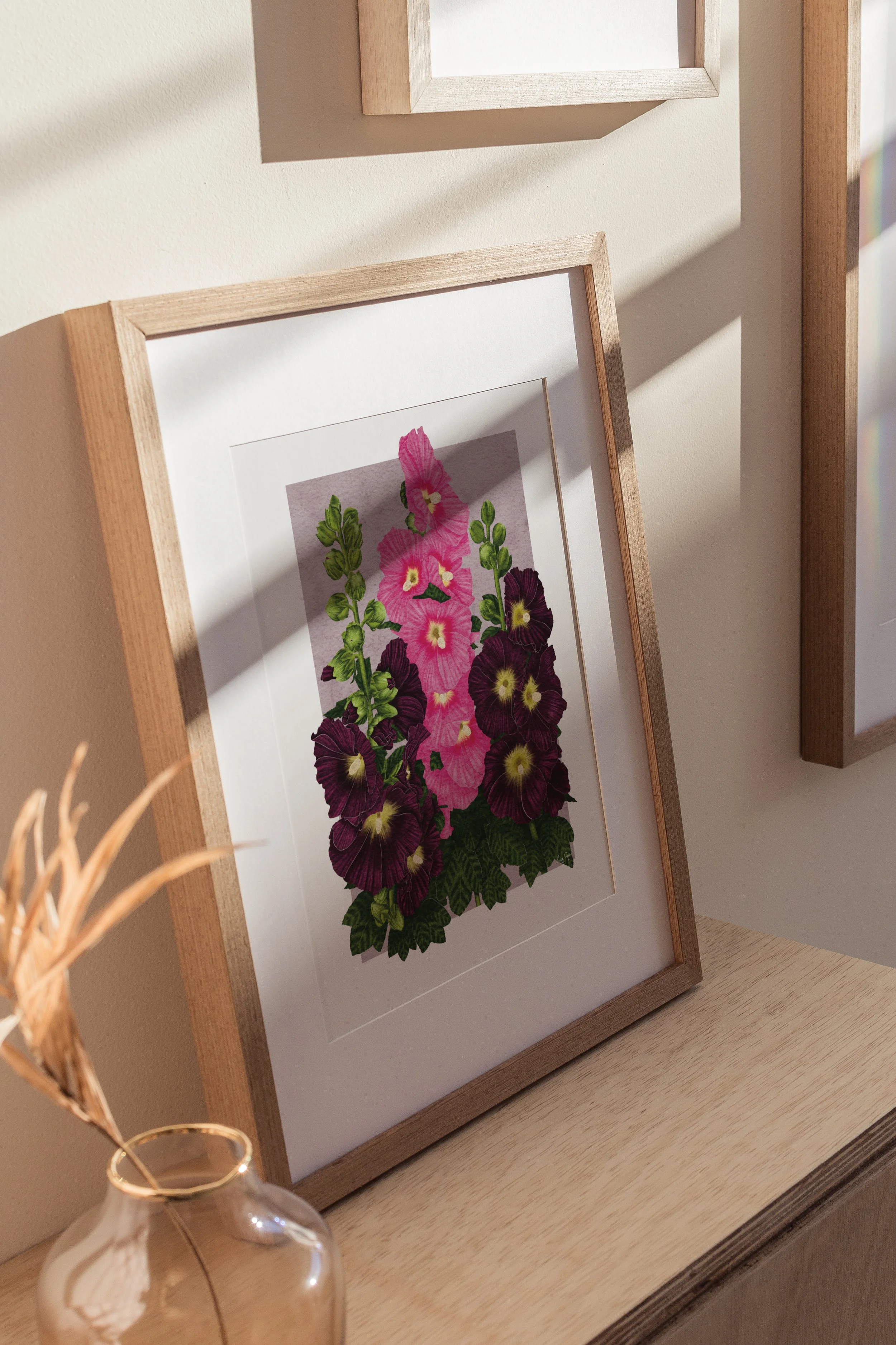 Framed Hollyhocks fine art print by Liz Govier Studio leaning against a wall in natural light with oak furniture and dried stems.