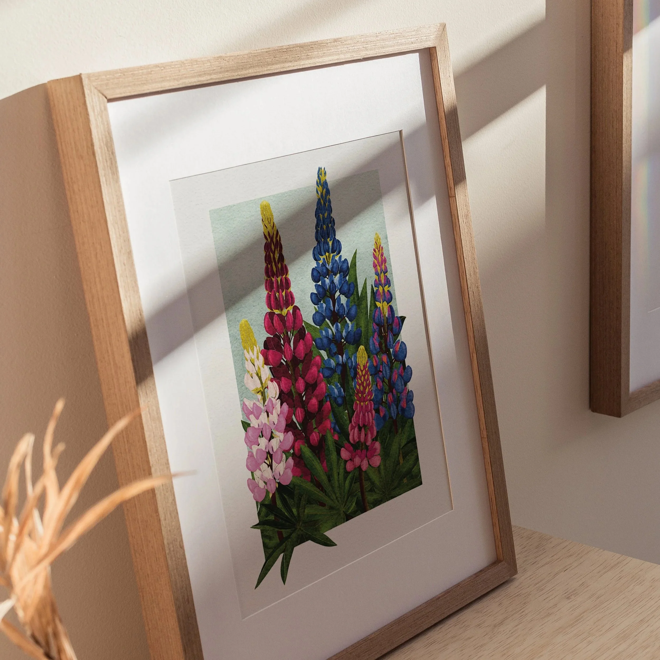 Close-up of the Lupins art print framed in light wood, showing vibrant pink, red, and blue lupin flowers against a soft green background, fine art botanical illustration.