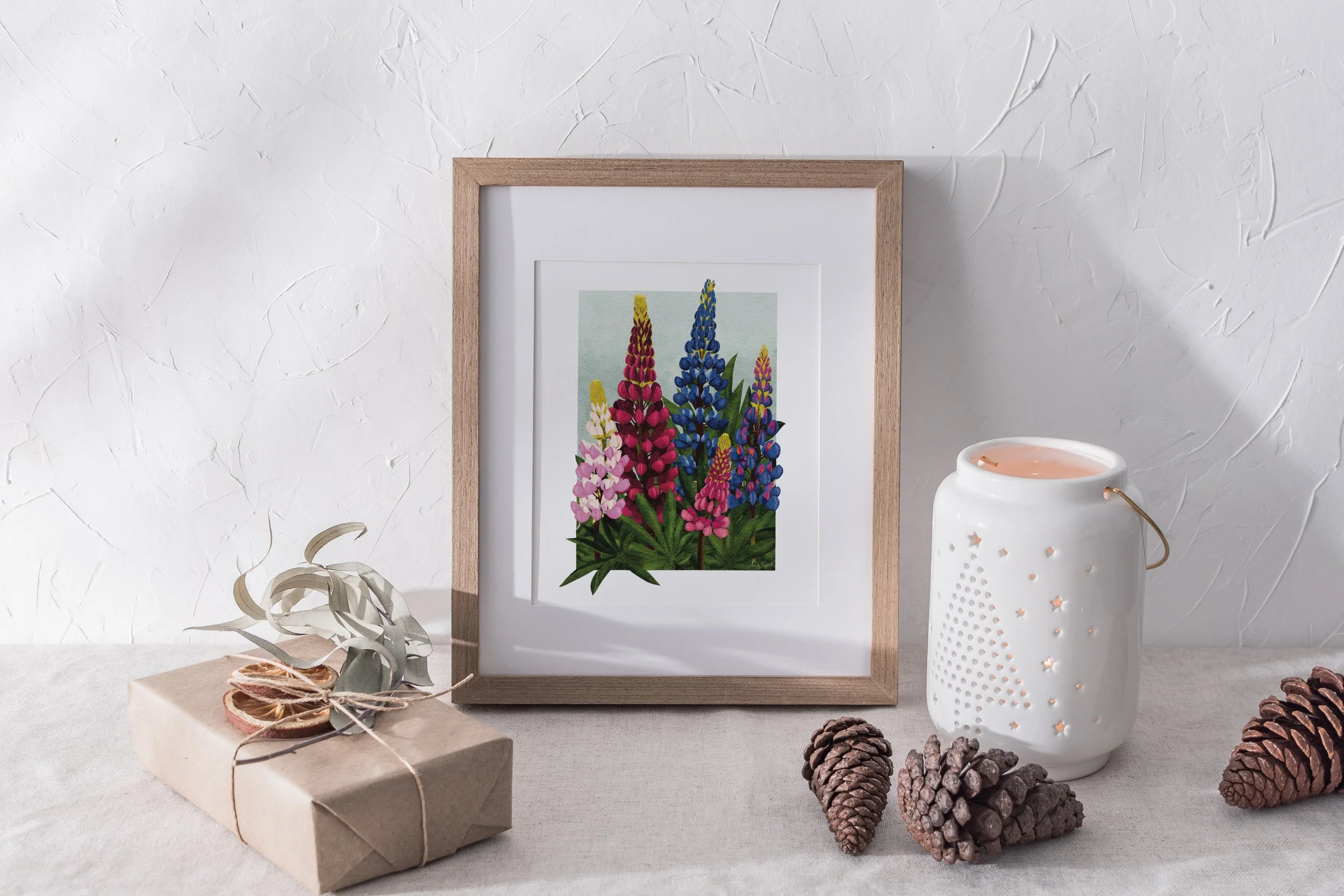 Framed Lupins fine art print styled with a kraft-paper gift, dried citrus, and a white lantern, arranged on a linen table.