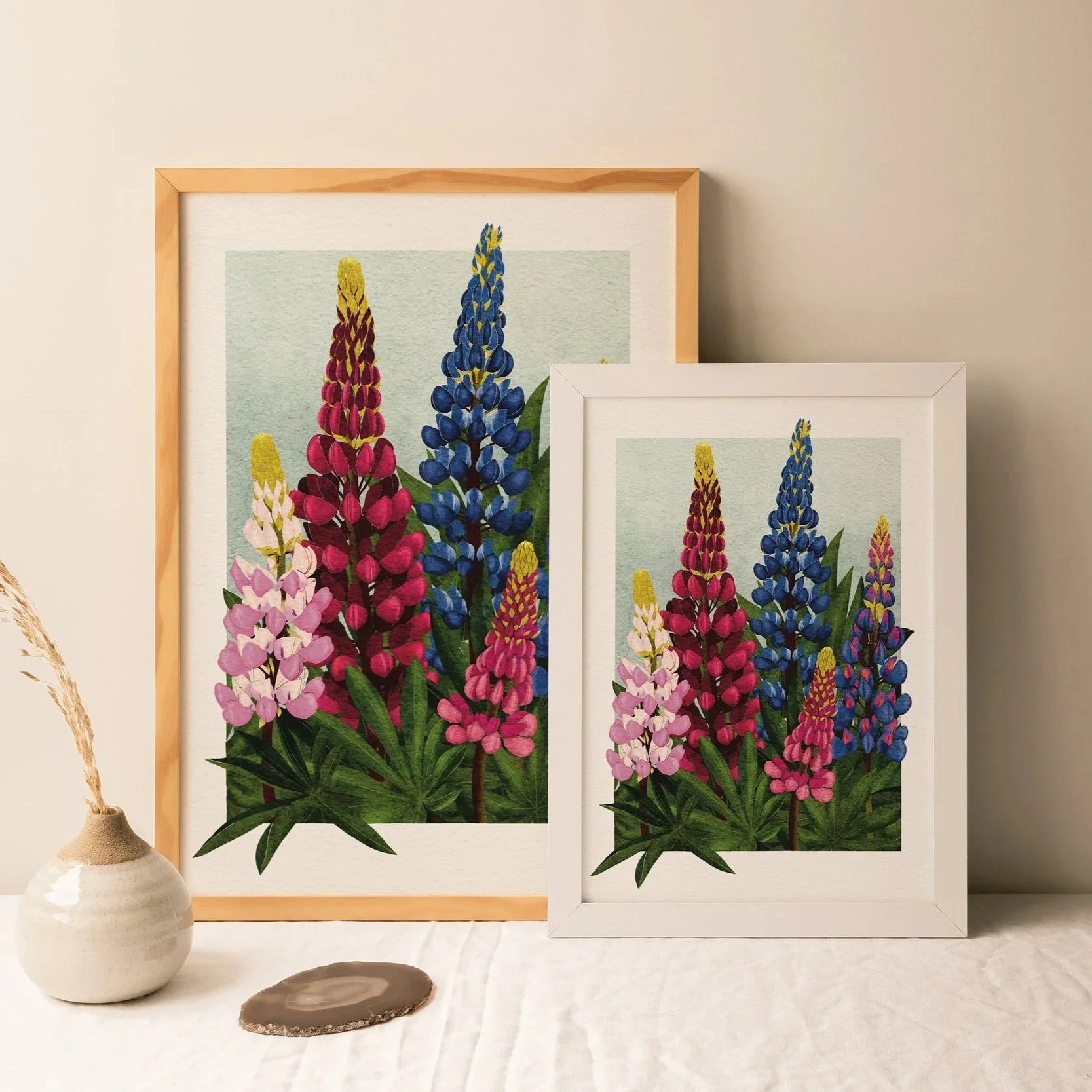 Two framed Lupins art prints in white and light wood frames, displayed side by side on a table with a ceramic vase, botanical wall art by Liz Govier Art Studio.