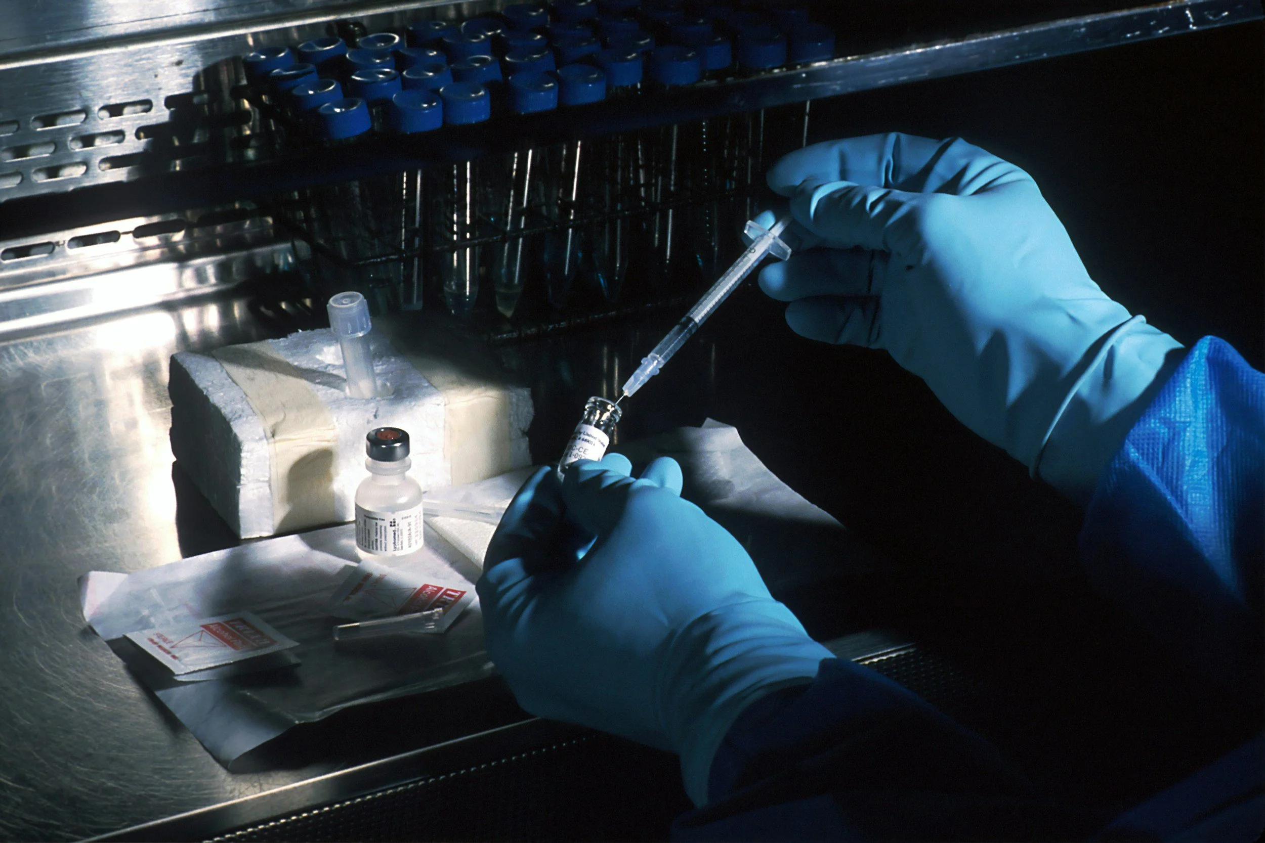 A scientist wearing blue gloves preparing a vaccine or medication with a syringe in a laboratory setting, surrounded by vials and medical supplies.