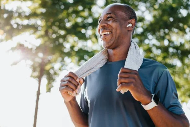 A man in athletic clothing smiling outdoors with a towel around his neck after exercising.