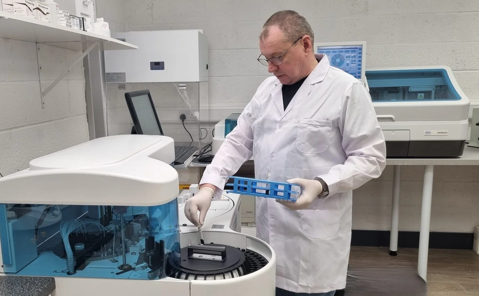 A scientist in a white lab coat and gloves operating a laboratory machine, with computer monitors and laboratory equipment in the background.