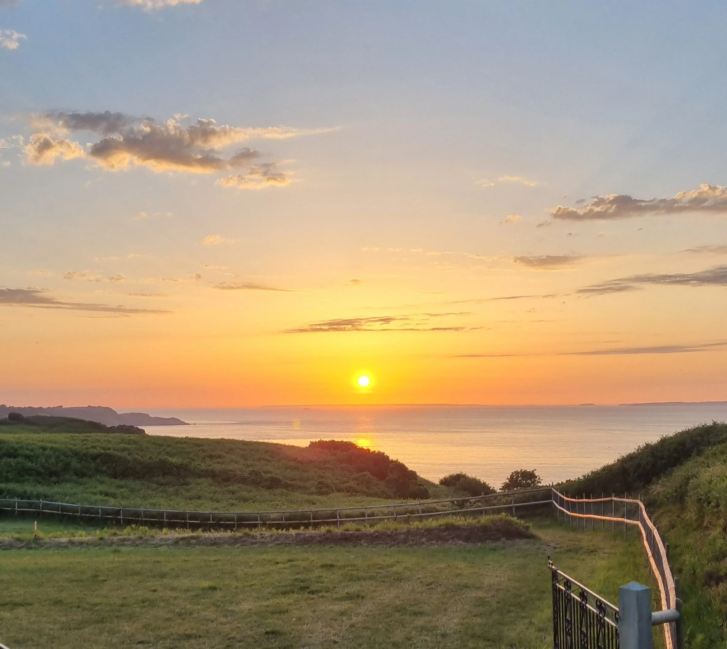 Sunset over the ocean with a grassy foreground, some bushes, a fence, and a partly cloudy sky.