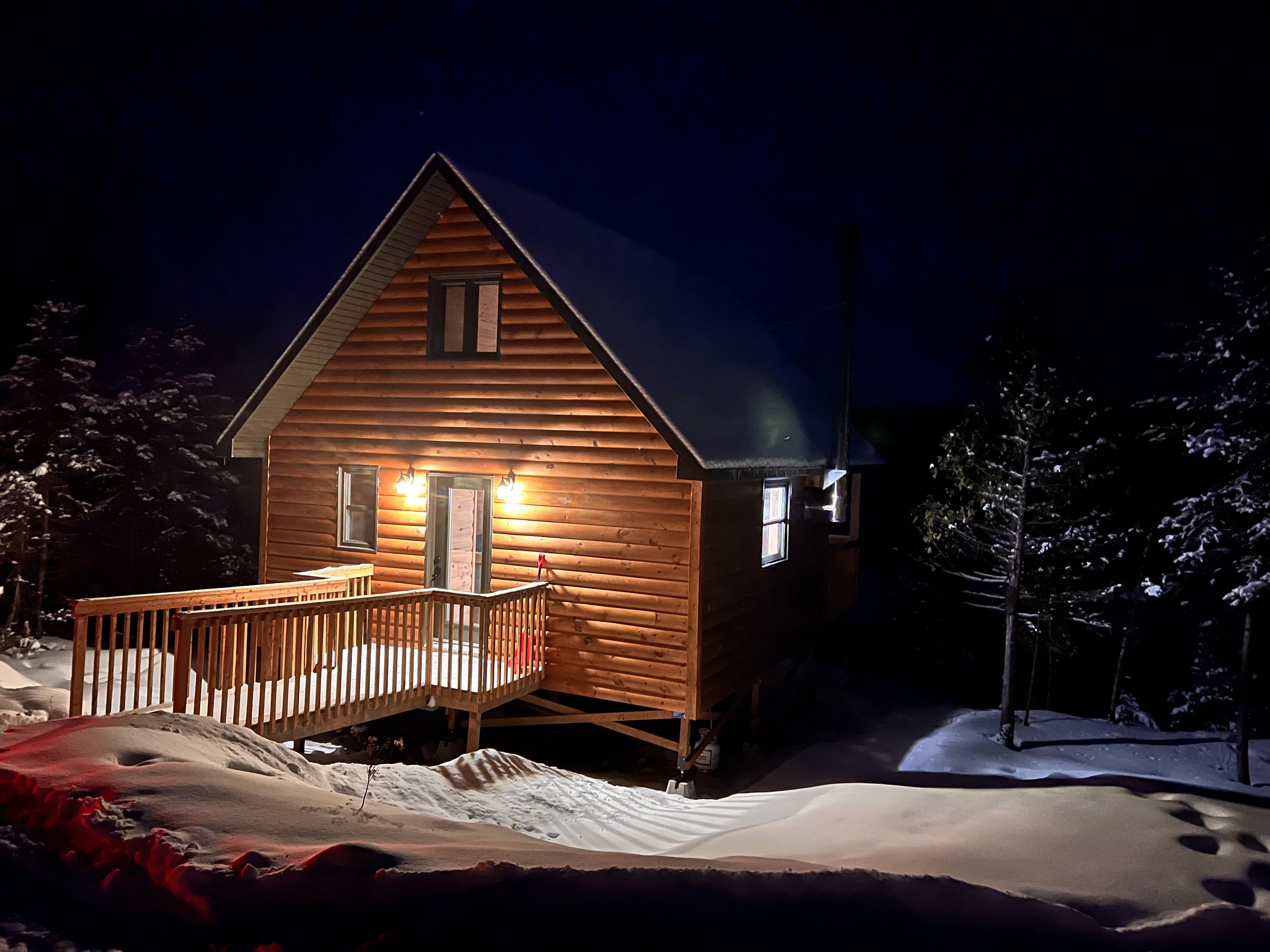 Cabane en bois dans la neige la nuit, éclairée par des lumières extérieures, entourée d'arbres enneigés.