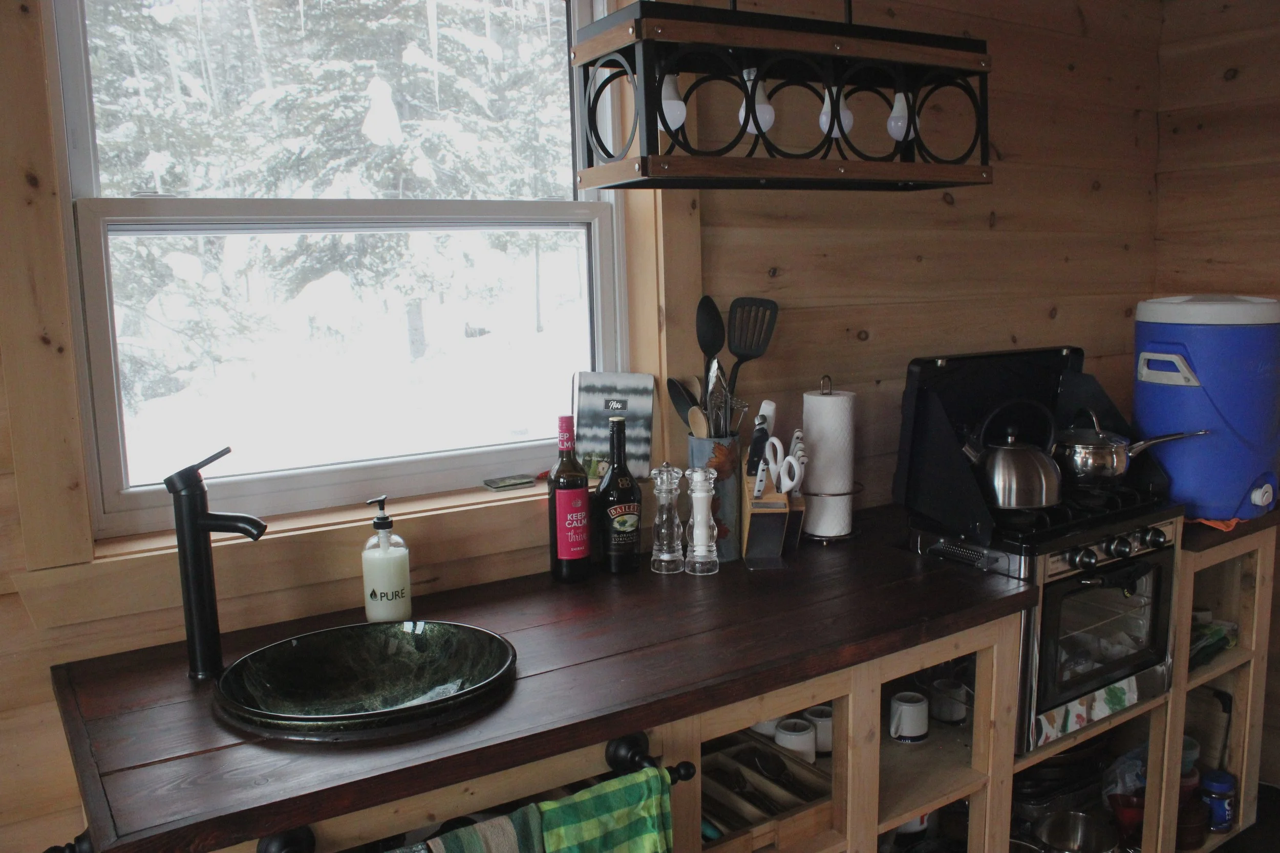 Intérieur d'une cuisine en bois avec une fenêtre donnant sur un paysage enneigé, un évier, différentes bouteilles, ustensiles, un film et un savon près du lavabo, ainsi qu'une unité de rangement en dessous.