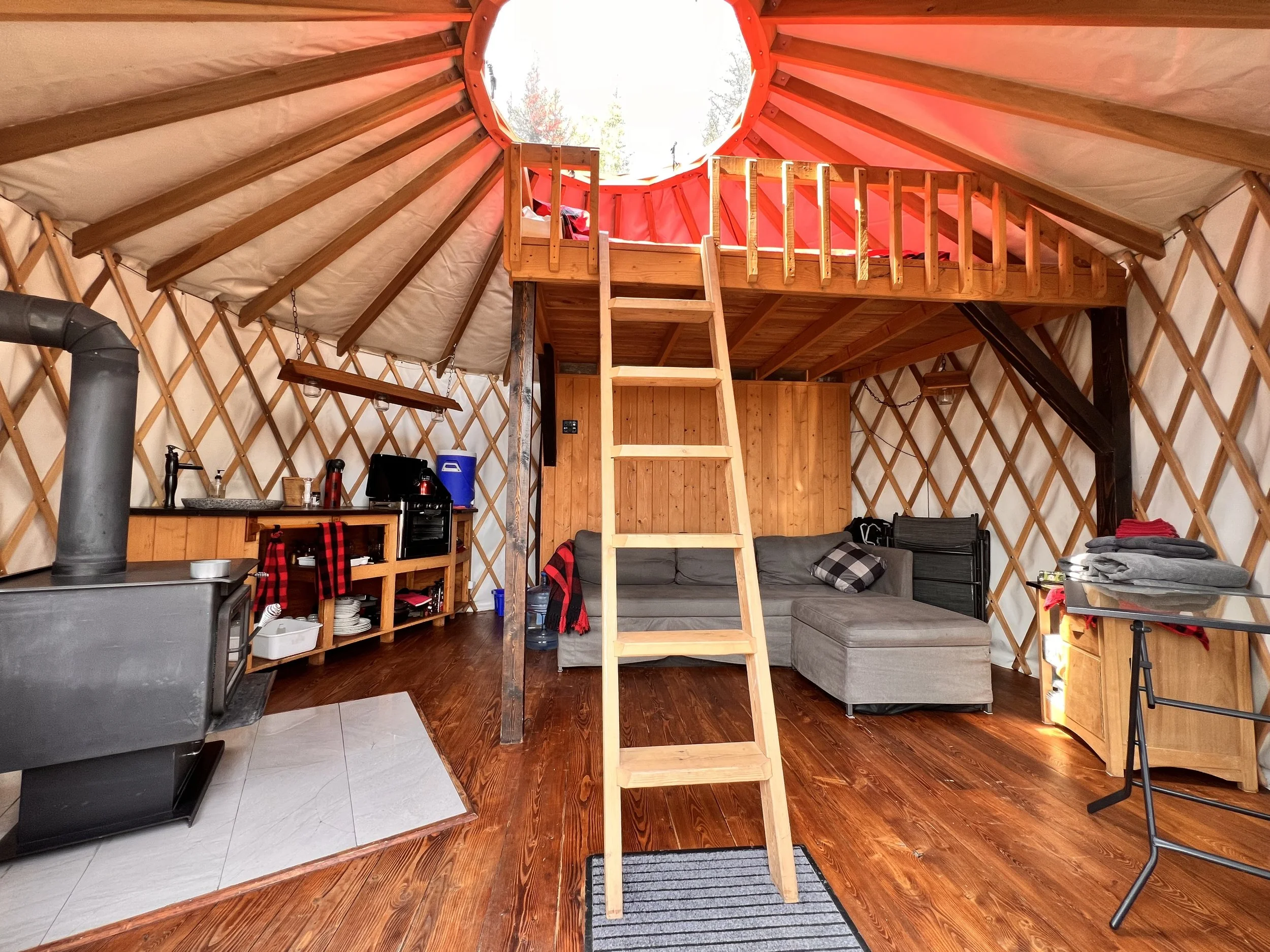 Intérieur d'une yourte avec un loft, un canapé en coin, une kitchenette avec un poêle, une table pliable et un tapis.