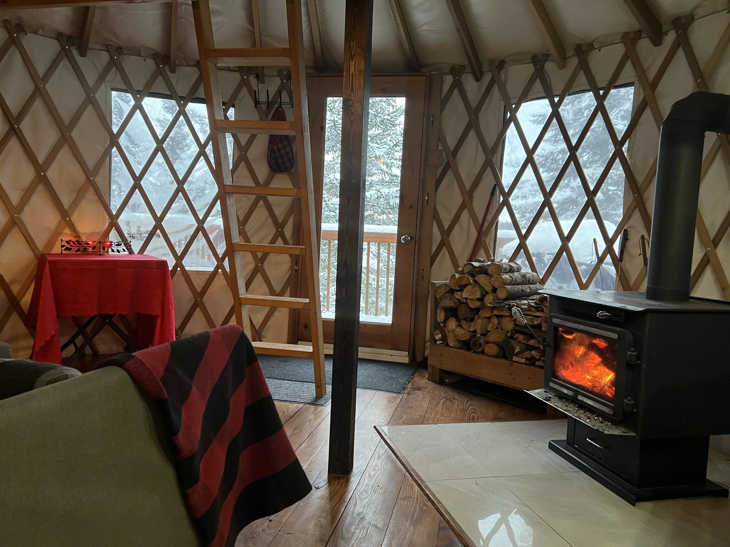 Intérieur d'une cabane en bois avec des fenêtres enneigées, un poêle à bois allumé, un tas de bois empilé, et une télévision, décorée pour l'hiver.