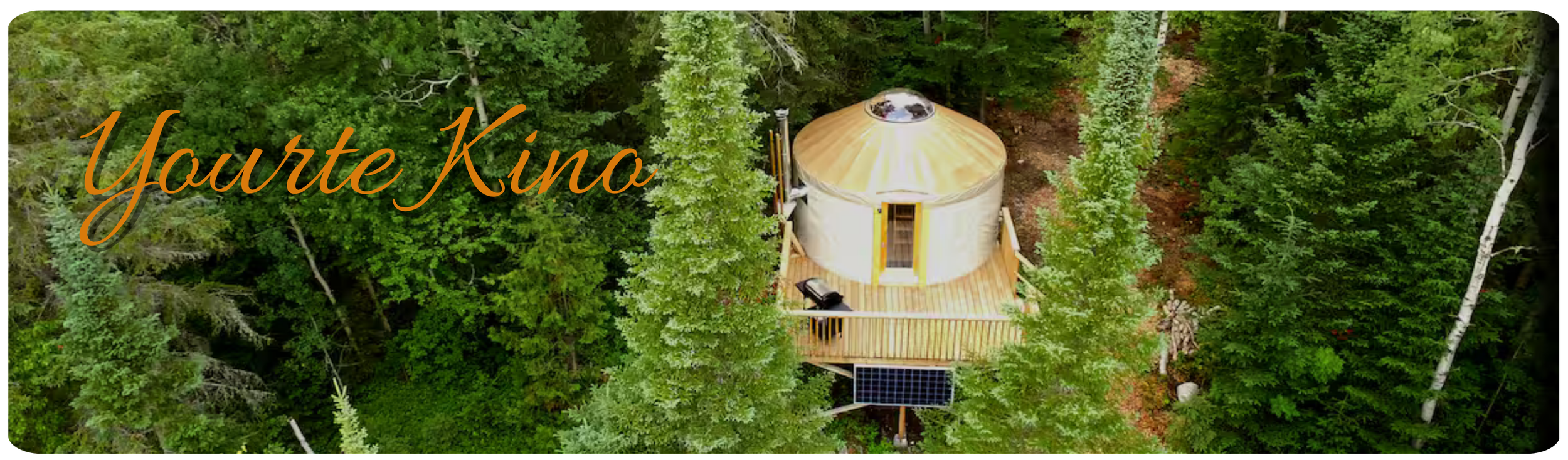 Une tiny house ronde en bois dans une forêt dense vue d'en haut, avec un toit en métal argenté, une terrasse en bois et une petite éolienne solaire.