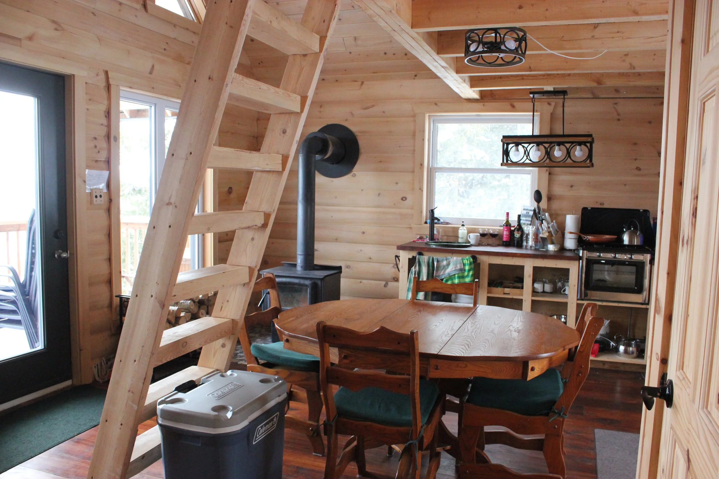 Intérieur d'une maison en bois avec escaliers en bois non fini, table en bois avec chaises, cheminée à bois, fenêtres laissant entrer la lumière, et cuisine sommaire avec quelques ustensiles.