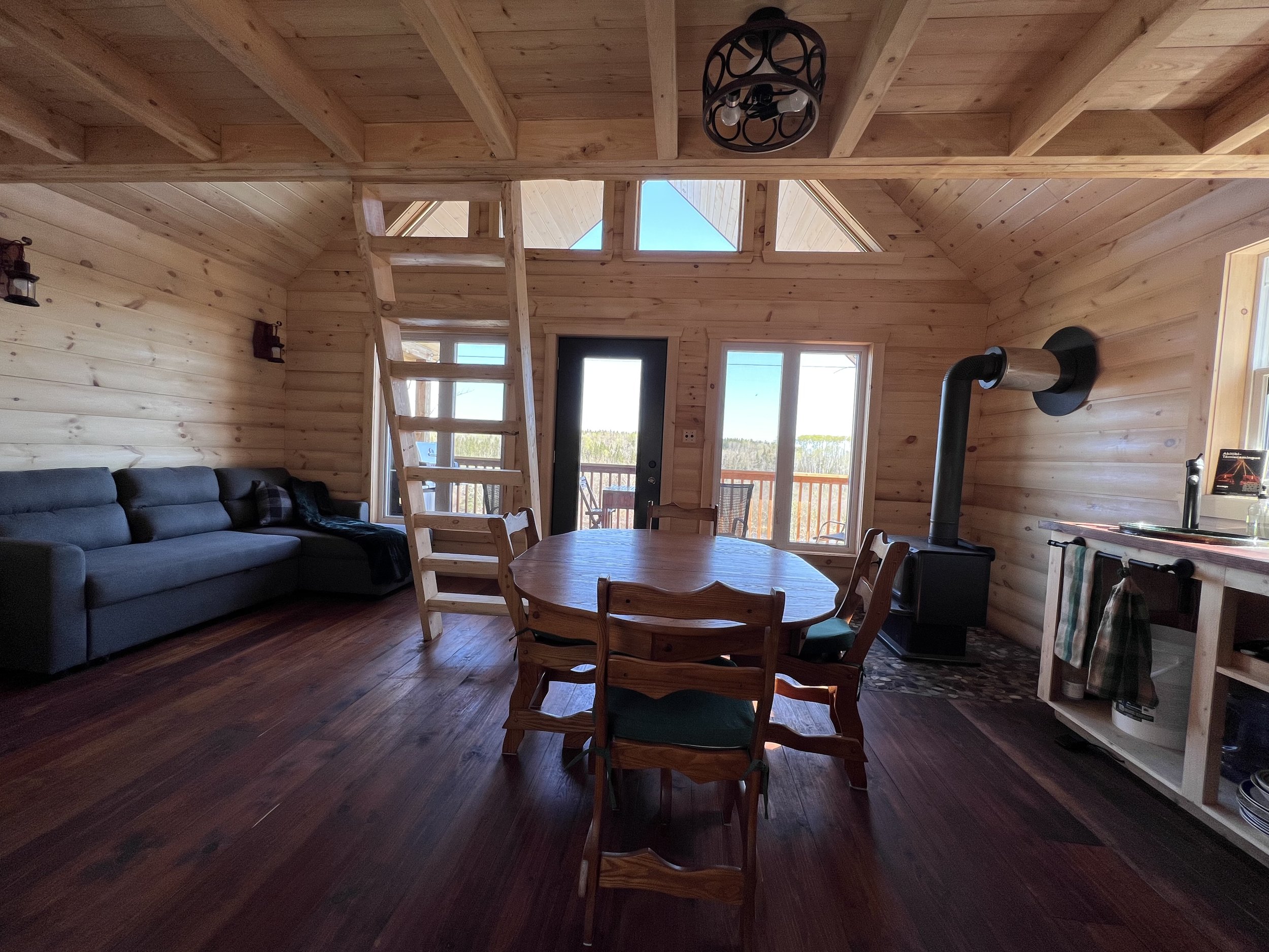 Intérieur d'une cabine en bois avec un salon, une table ronde en bois avec des chaises, un poêle à bois, une cuisine partielle, et un escalier en bois menant à un mezzanine. De grandes fenêtres laissent entrer la lumière naturelle.