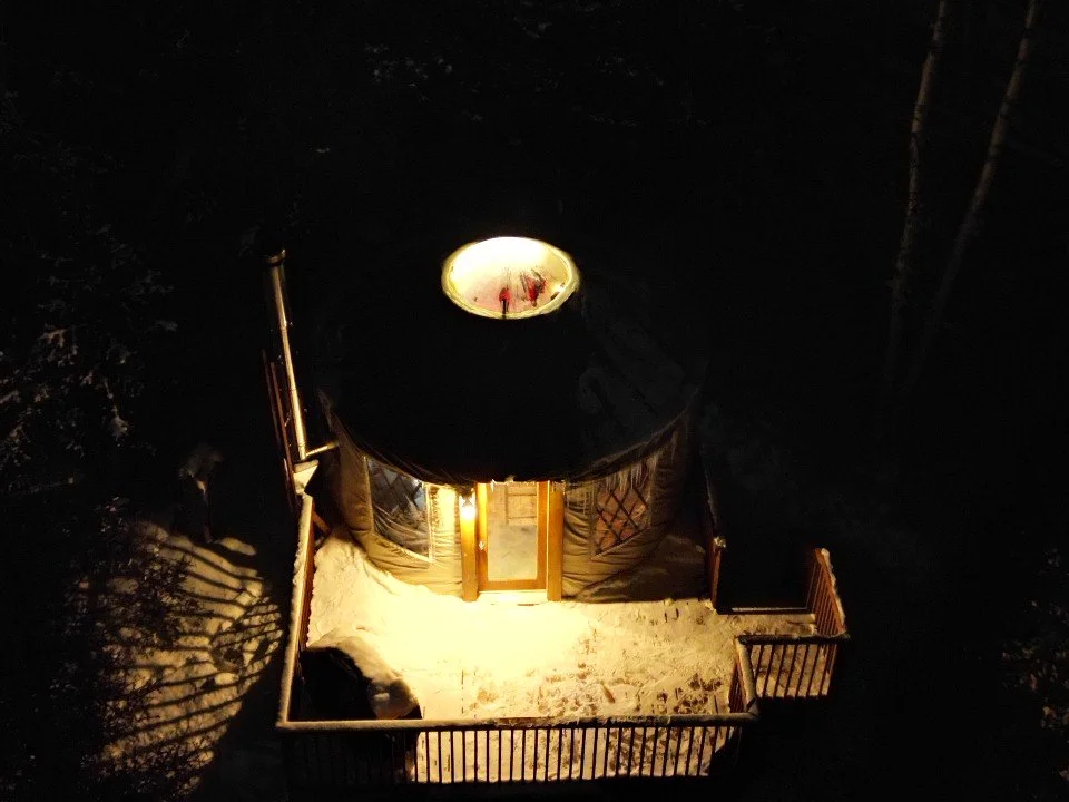 Une cabane en bois avec un toit noir, éclairée à l'intérieur, située dans un environnement forestier nocturne. La cabane a un balconvade, et il y a une fenêtre ronde au sommet du toit reflétant deux personnes.