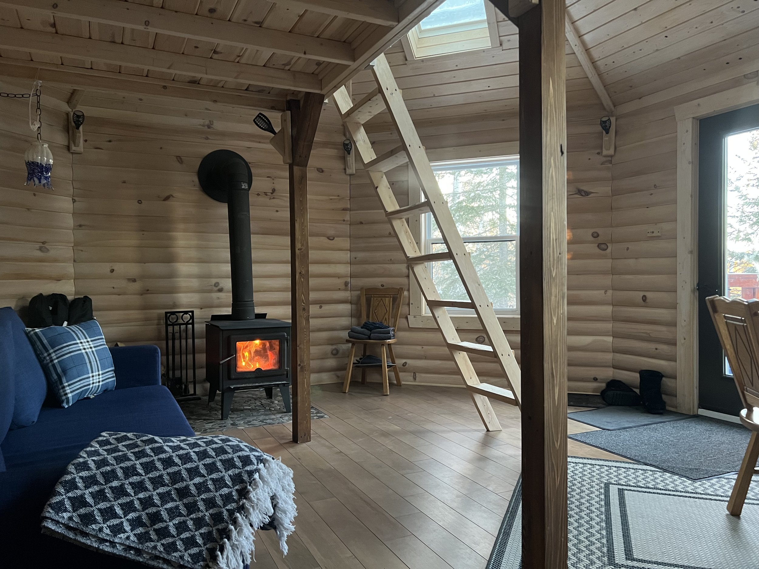 Intérieur d'une cabine en bois avec un poêle à bois, une échelle menant à un espace en mezzanine, des fenêtres laissant entrer la lumière naturelle et des meubles rustiques.