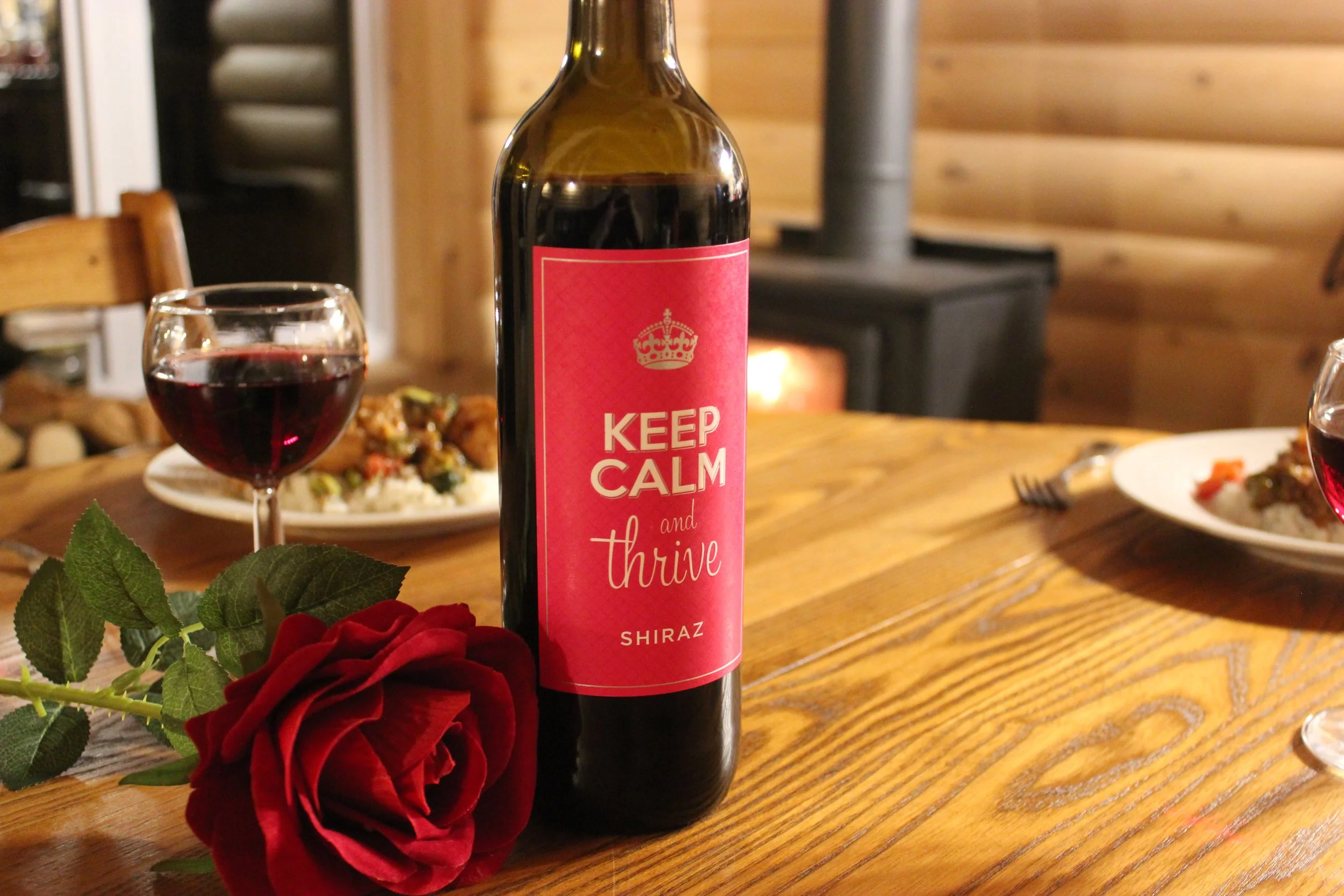 Une bouteille de vin rouge avec une étiquette rose disant « Keep Calm and Thrive Shiraz » sur une table en bois, accompagnée de deux verres de vin rouge, une assiette de nourriture, une rose rouge et un feu de cheminée en arrière-plan.