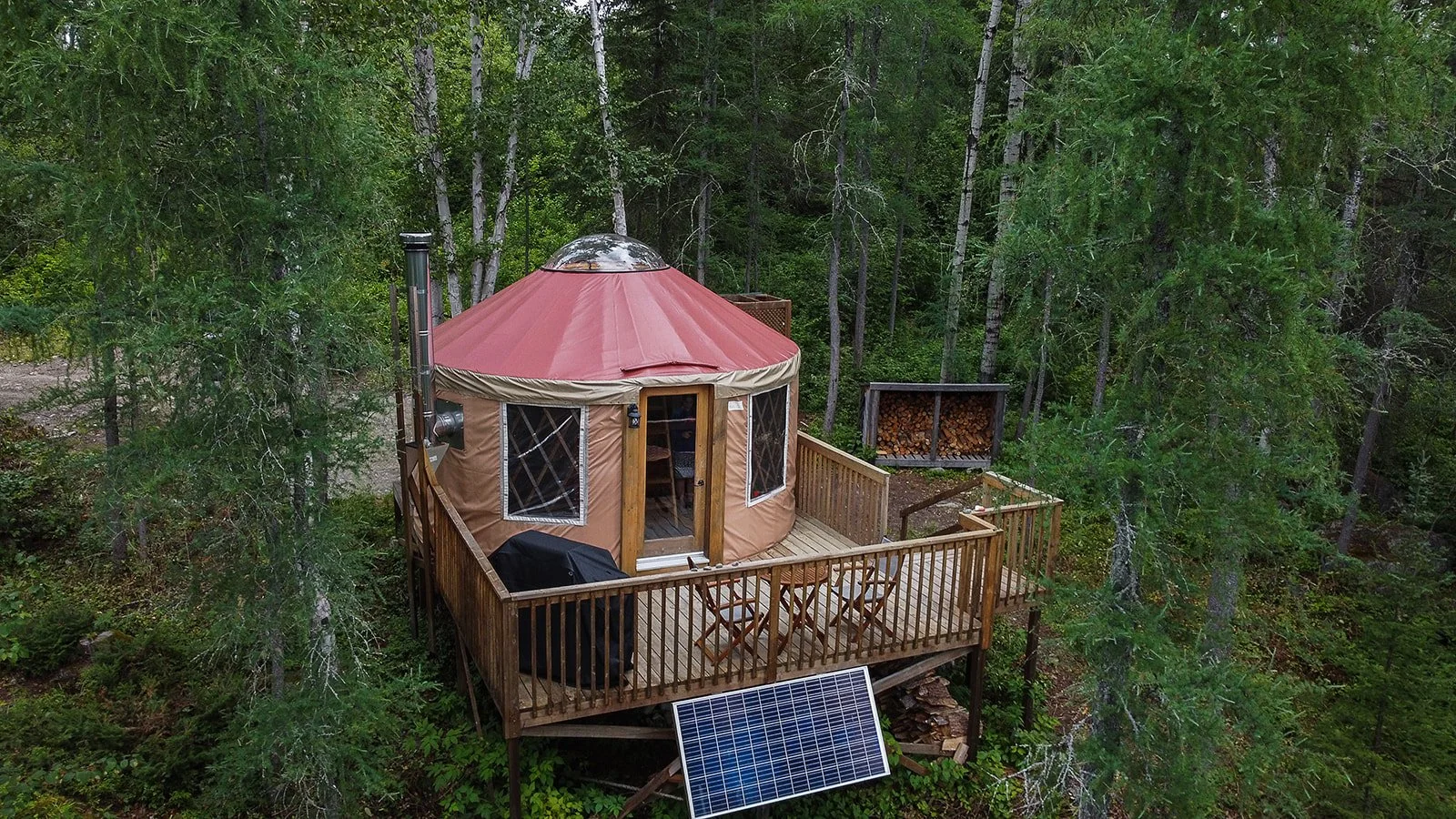 Une yourte en bois et toile dans une forêt, avec une terrasse équipée d'une table et de chaises, panneau solaire en dessous, stockage de bois à l'arrière.