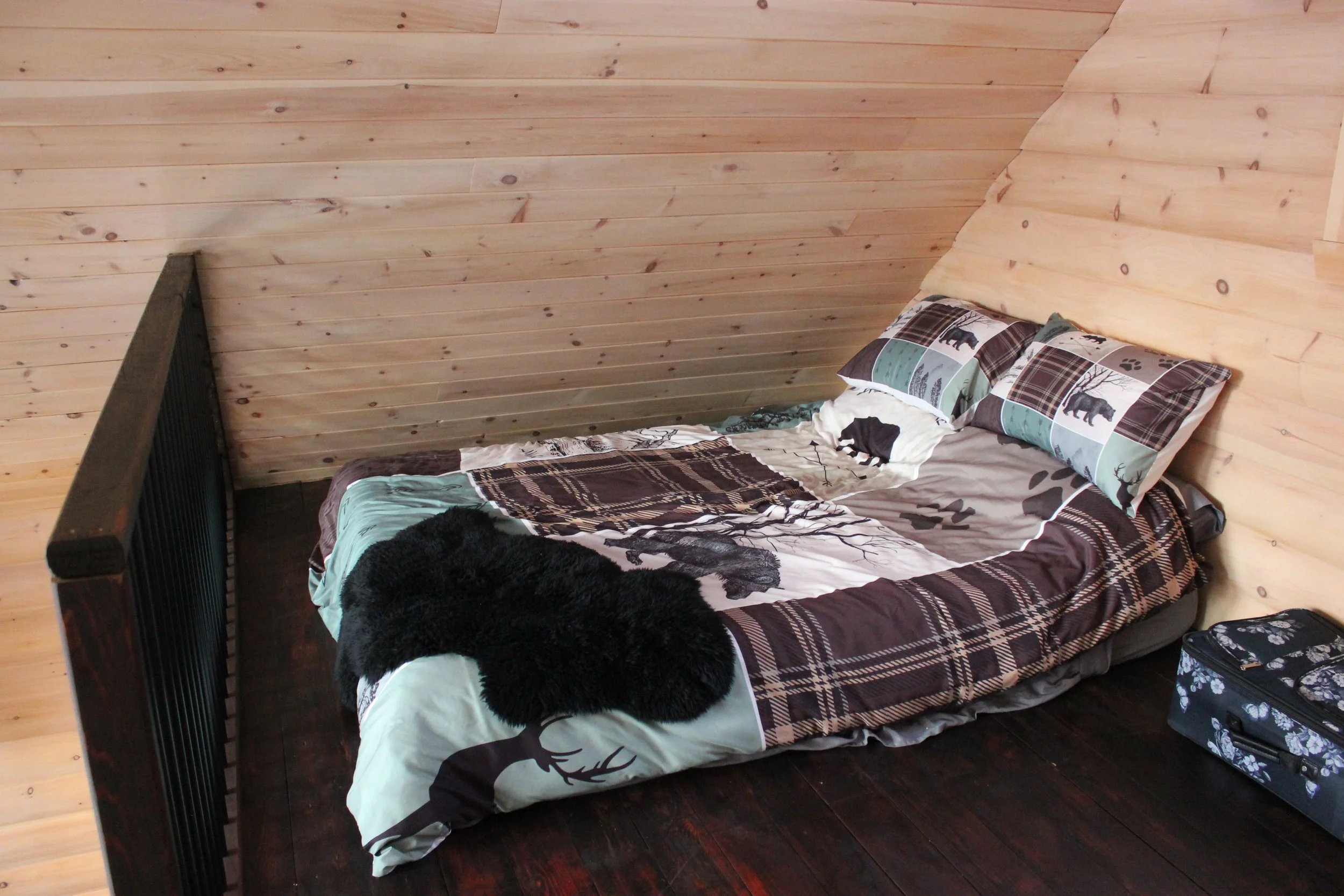 Lit avec une couette à motif forestier, deux oreillers, un tapis en fausse fourrure noire, une valise avec des fleurs sur la droite, dans une chambre en bois.