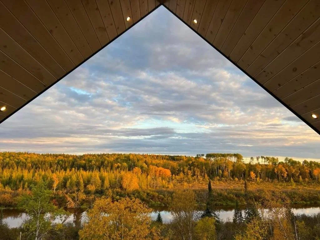 Vue sur la nature avec des arbres aux couleurs automnales, un lac et un ciel nuageux, vue depuis une structure en bois avec un toit en triangle.