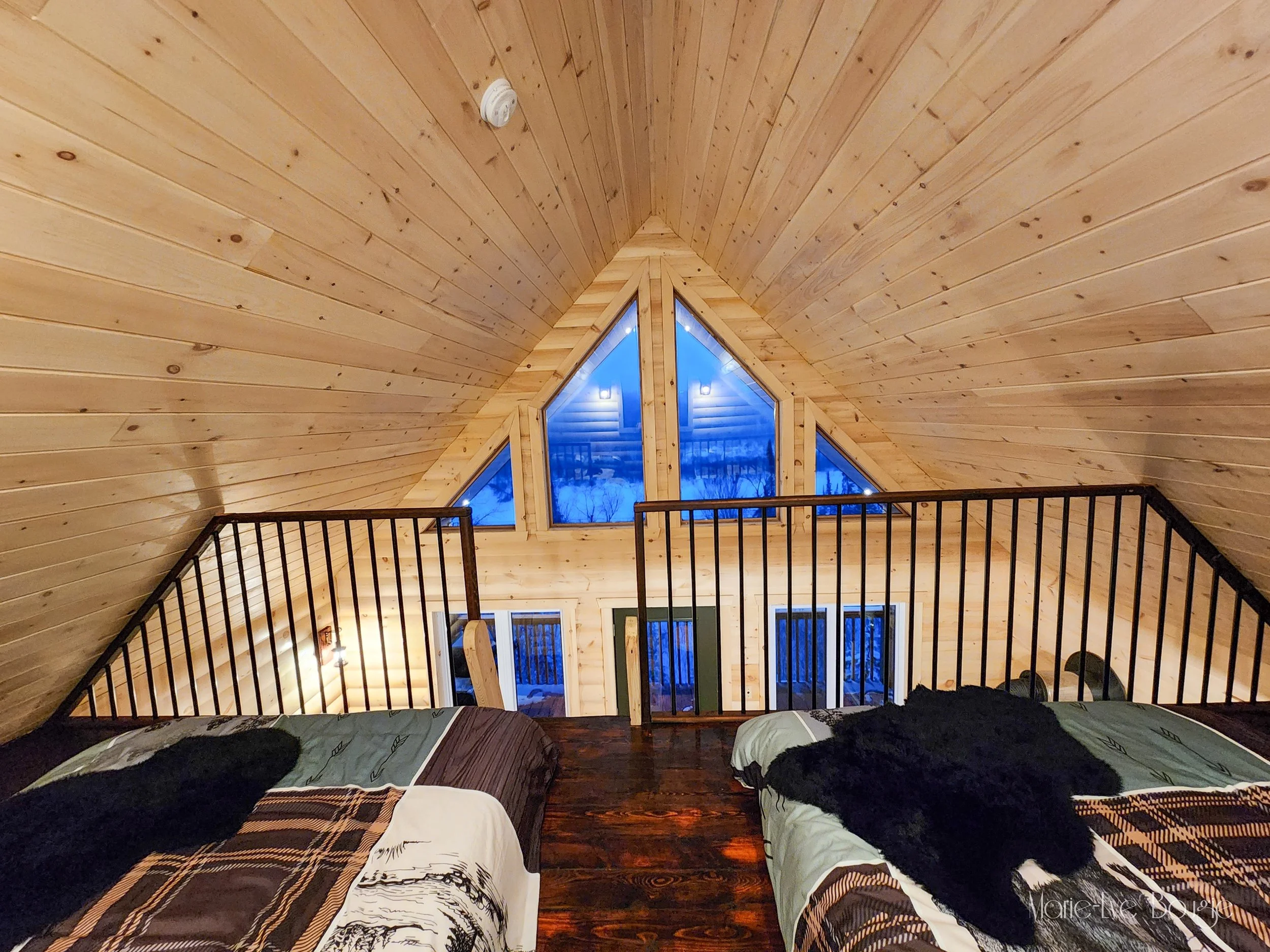 Intérieur d'une chambre dans un chalet en bois avec deux lits, une rambarde en métal, une grande fenêtre avec vue sur la forêt enneigée et un ciel bleu, et un plafond en bois pointu.