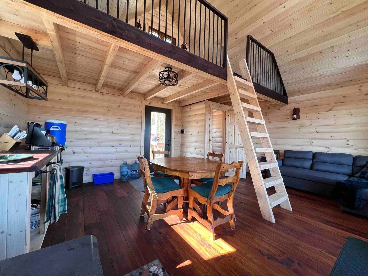 Intérieur d'une cabine en bois avec un salon, une table ronde en bois, quatre chaises, un canapé noir, une échelle menant à un loft, et une cuisine partielle.