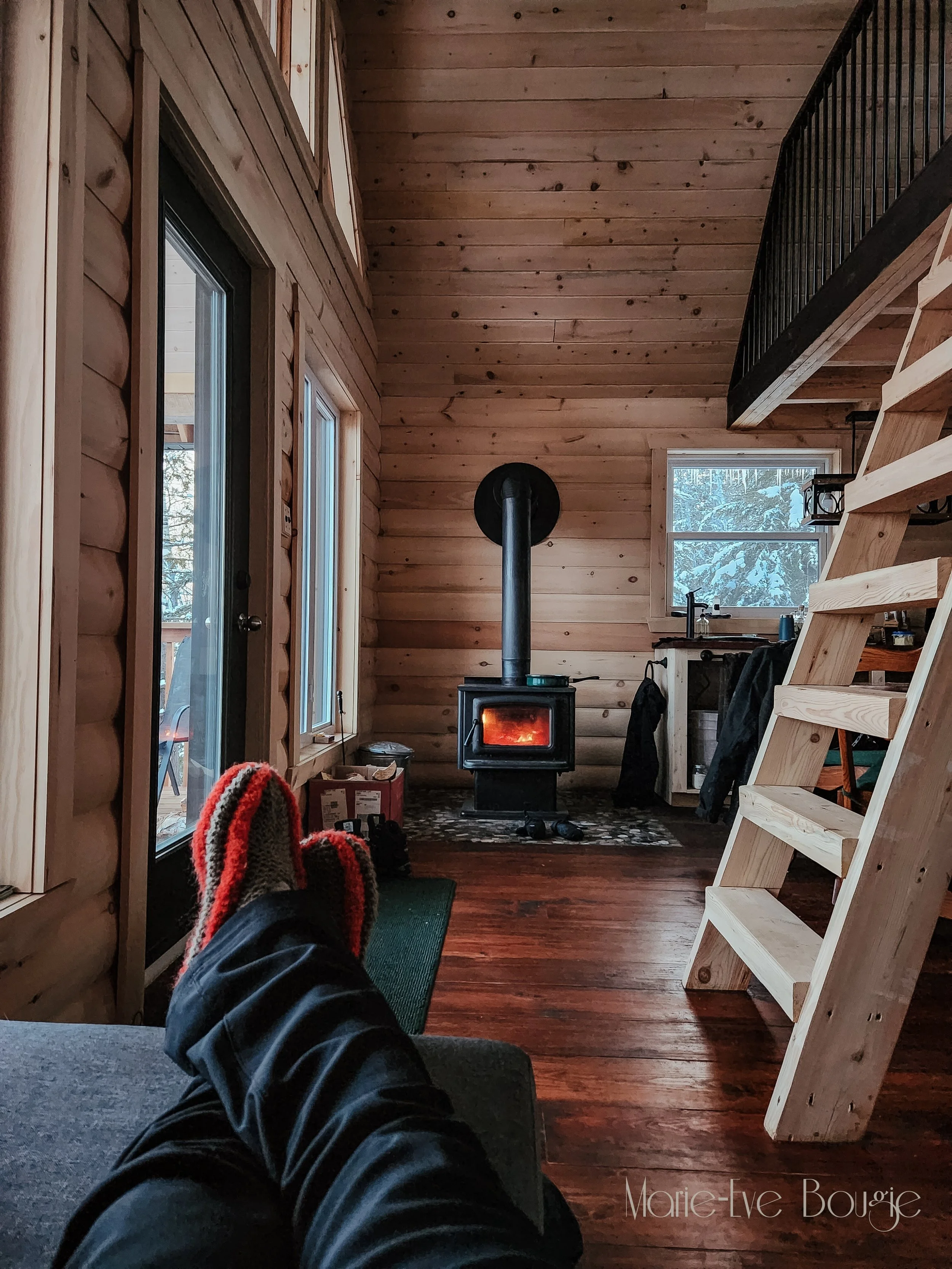 Intérieur d'une cabine en bois avec un poêle à bois allumé, une fenêtre montrant un paysage enneigé, un escalier en bois, et des jambes avec des chaussettes tricotées rouges, noires et gris posées sur un sofa.