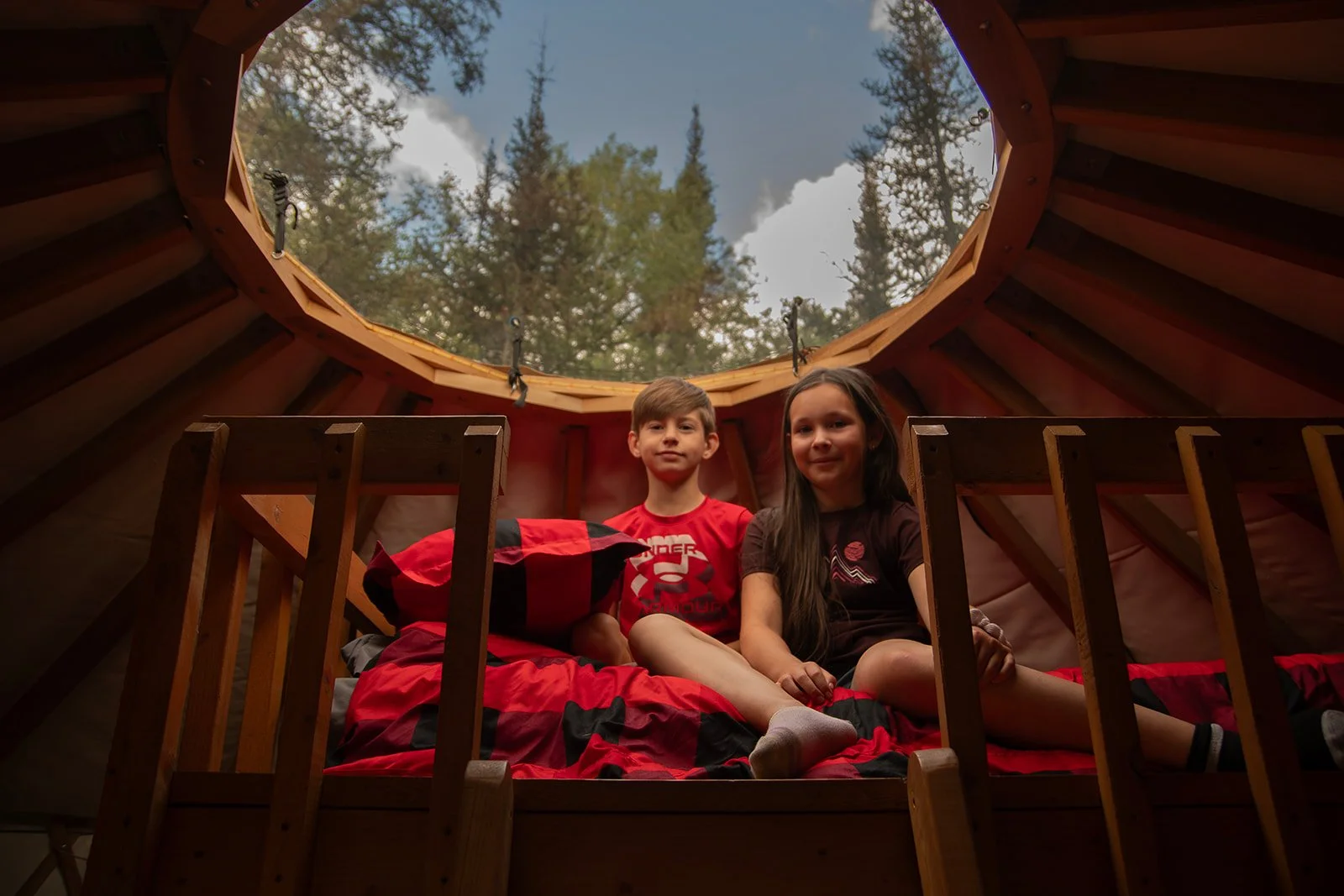 Deux enfants assis dans une cabane en bois, regardant par un toit ovale ouvert, avec des arbres et le ciel visible en arrière-plan.
