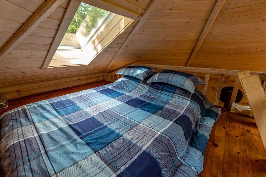 Lit dans une chambre mansardée avec un plafond en bois et un plafond en pente, sous une fenêtre de toit laissant entrer la lumière naturelle, avec des draps à carreaux bleus.