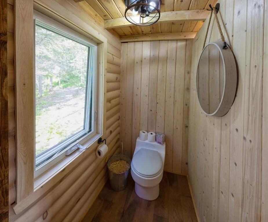 Petite salle de bain en bois avec un lavabo, un miroir rond suspendu, un rouleau de papier toilette, une fenêtre laissant entrer la lumière naturelle, et un plafond en bois avec un lustre noir.