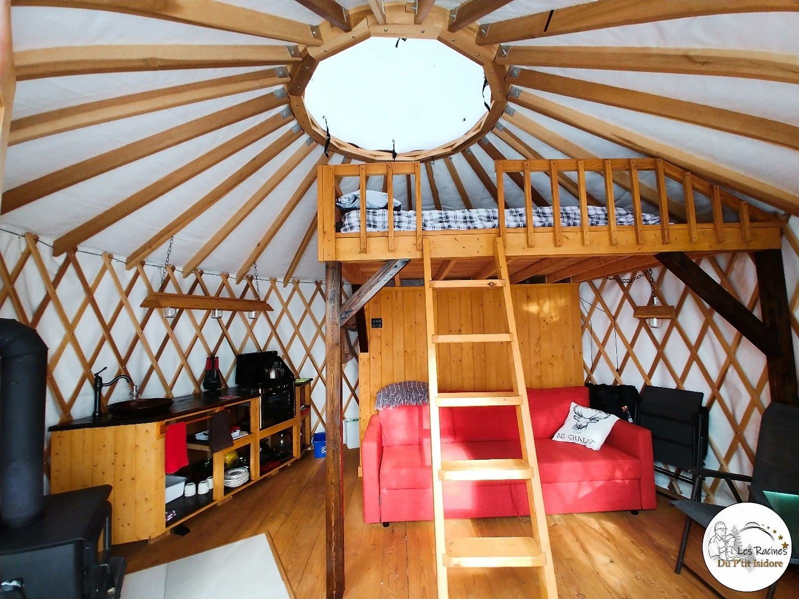 Intérieur d'une yourte avec un lit en mezzanine, un canapé rouge, une cuisine compacte, et des éléments en bois, éclairée par une lumière naturelle venant d'une ouverture en forme d'octogone au plafond.