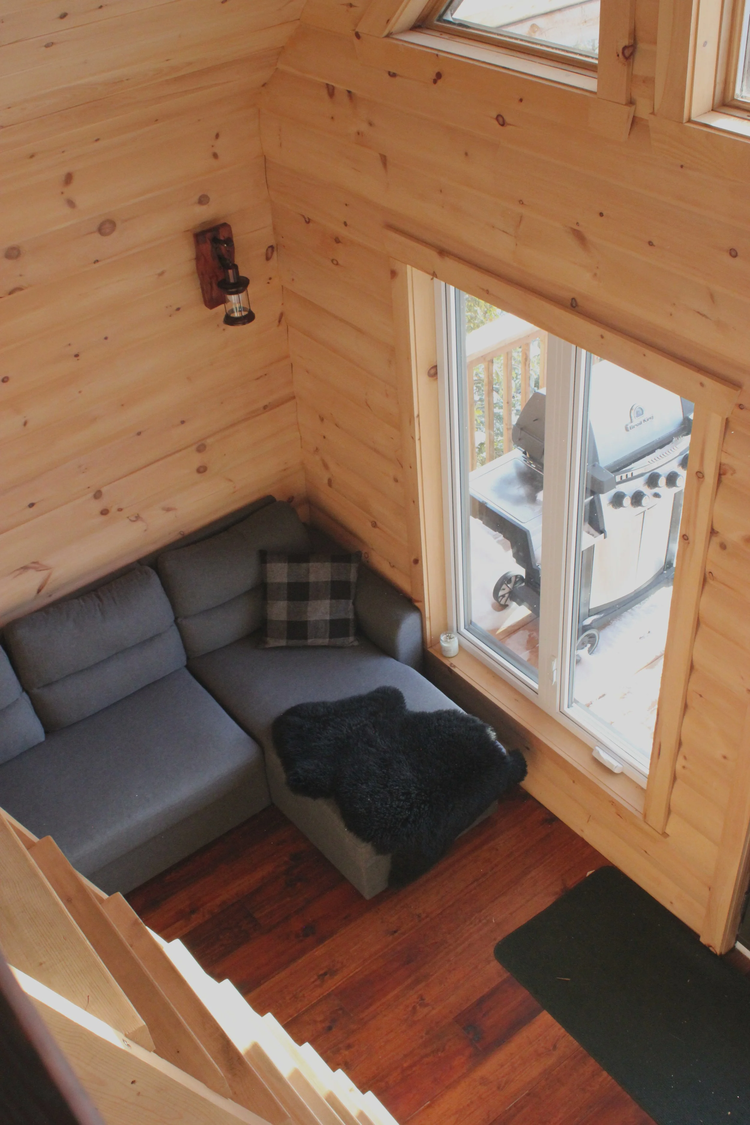 Salon avec un canapé gris, un coussin à carreaux, une couverture en fourrure noire, un mur en bois et un barbecue visible par la fenêtre.