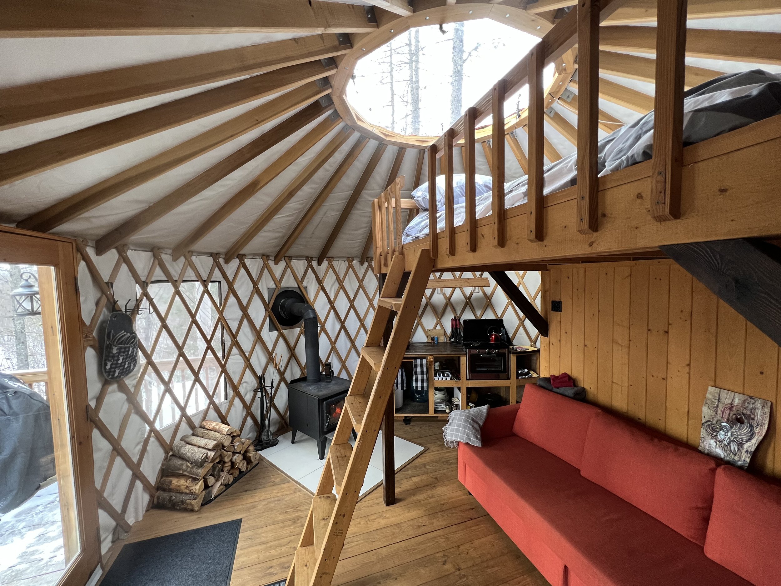 Un intérieur de cabine en bois avec un lit dans une mezzanine, une cheminée à bois, une pile de bois, une table avec une chaîne stéréo, un canapé rouge et des fenêtres donnant sur la neige et des arbres.