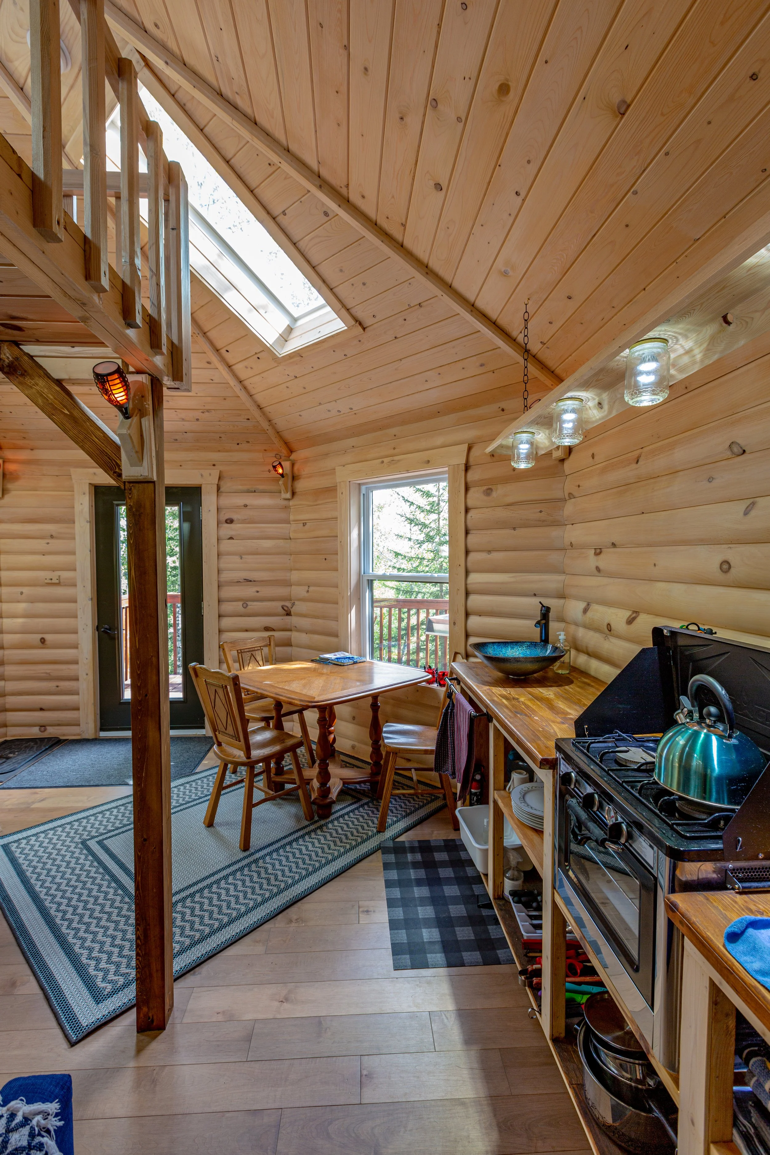 Intérieur d'une cuisine en bois avec une table en bois, des chaises, un poêle à gaz, et une fenêtre donnant sur l'extérieur. Un plafond en pan de bois avec lampes suspendues en bocaux et des lumières chauffantes.