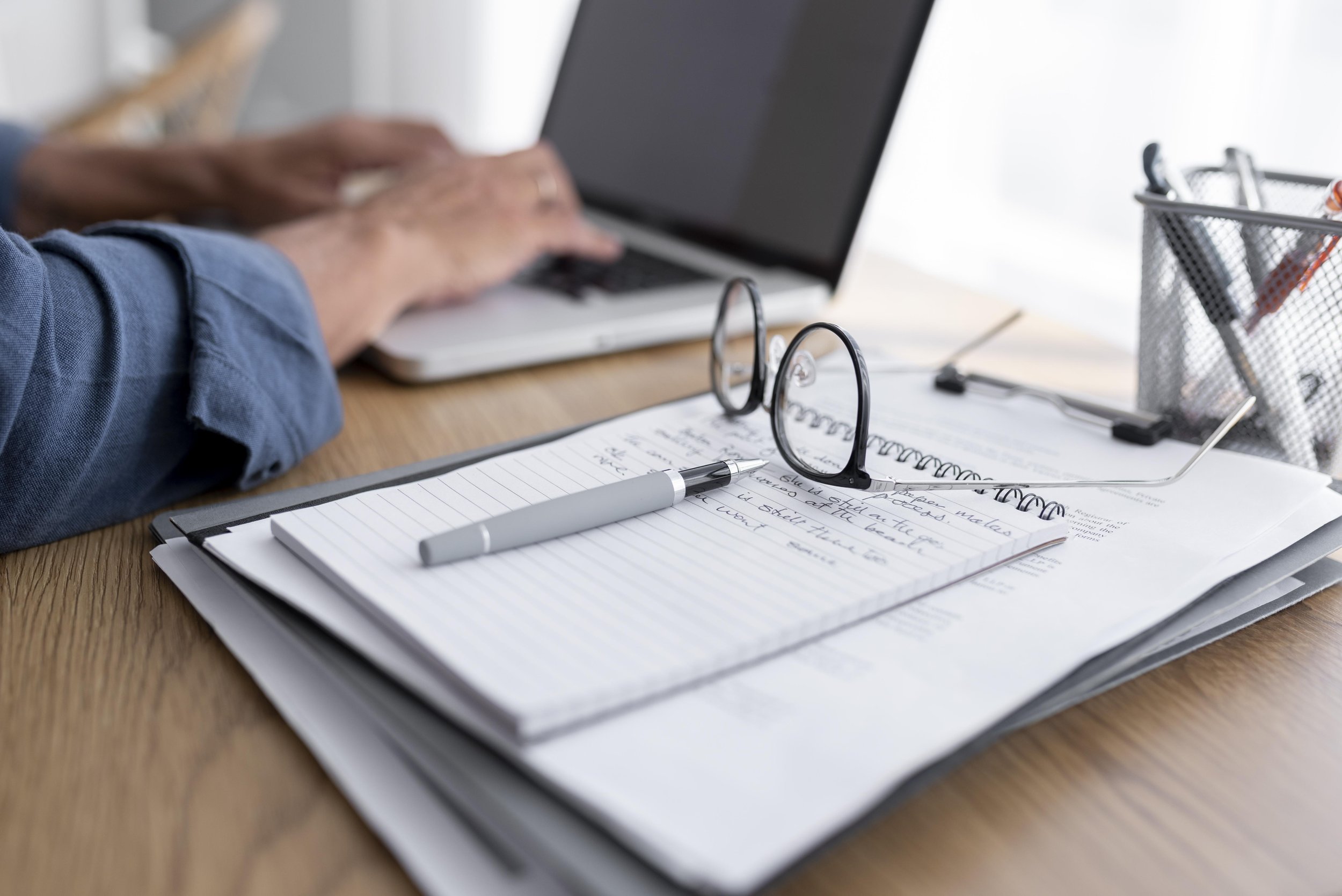 A desk with a notepad, glasses, pen, and papers, with a person using a laptop in the background.