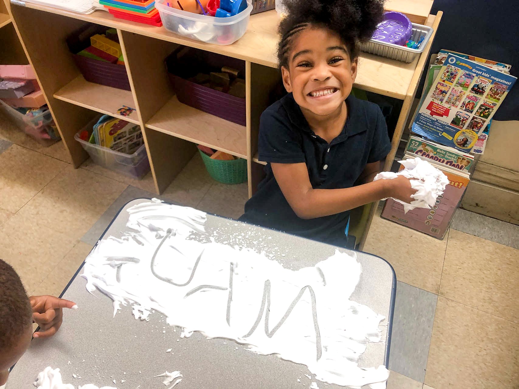 A young girl with a big smile, holding a cloth and sitting at a Tots Yearn to Learn classroom table with white foam on it, learn to write letters.