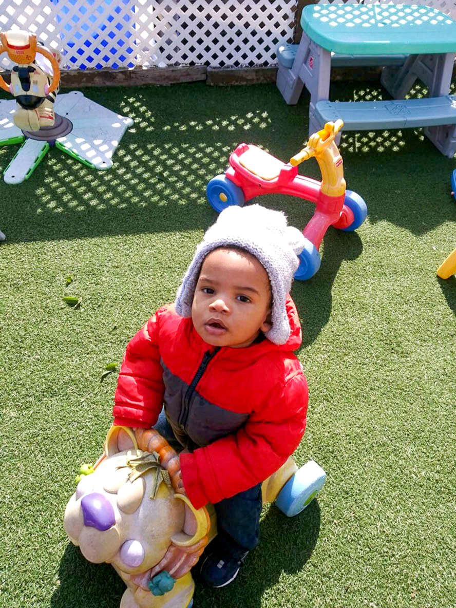 A young child wearing a gray hat and red jacket sitting on a small, colorful plastic ride-on toy with a lion face, outdoors at Tots Yearn to Learn on a grassy area with playground toys and a white lattice fence in the background.