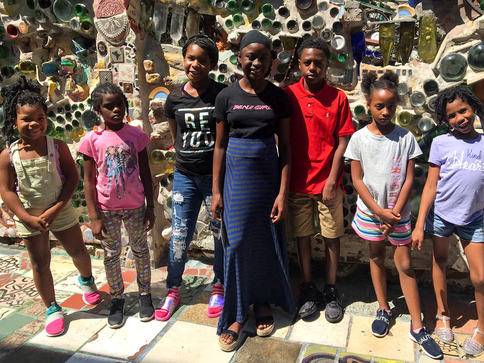A group of seven children and one adult standing in front of a colorful mosaic wall made of glass and other materials.