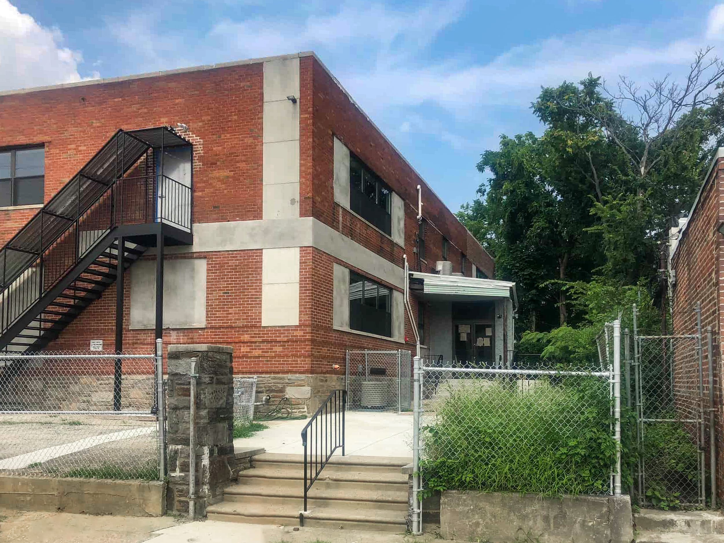 Tots Yearn to Learn Larchwood, a two-story brick building with an external metal staircase, surrounded by a fence, with some greenery and trees nearby under a partly cloudy sky.
