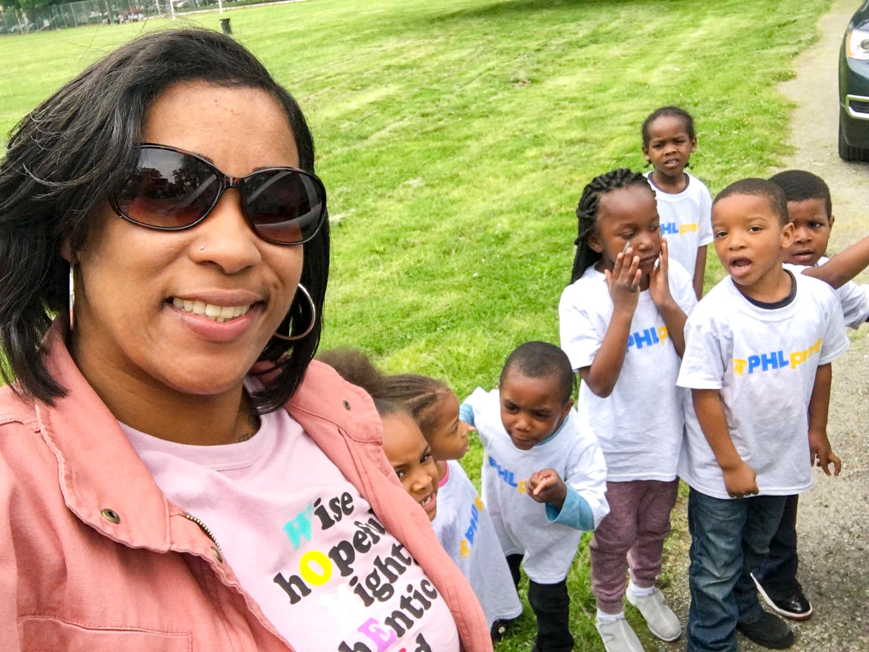 A Tots Yearn to Learn teacher taking a selfie outdoors with six young, excited-looking children in white T-shirts, on a grassy area with a car partially visible on the right.