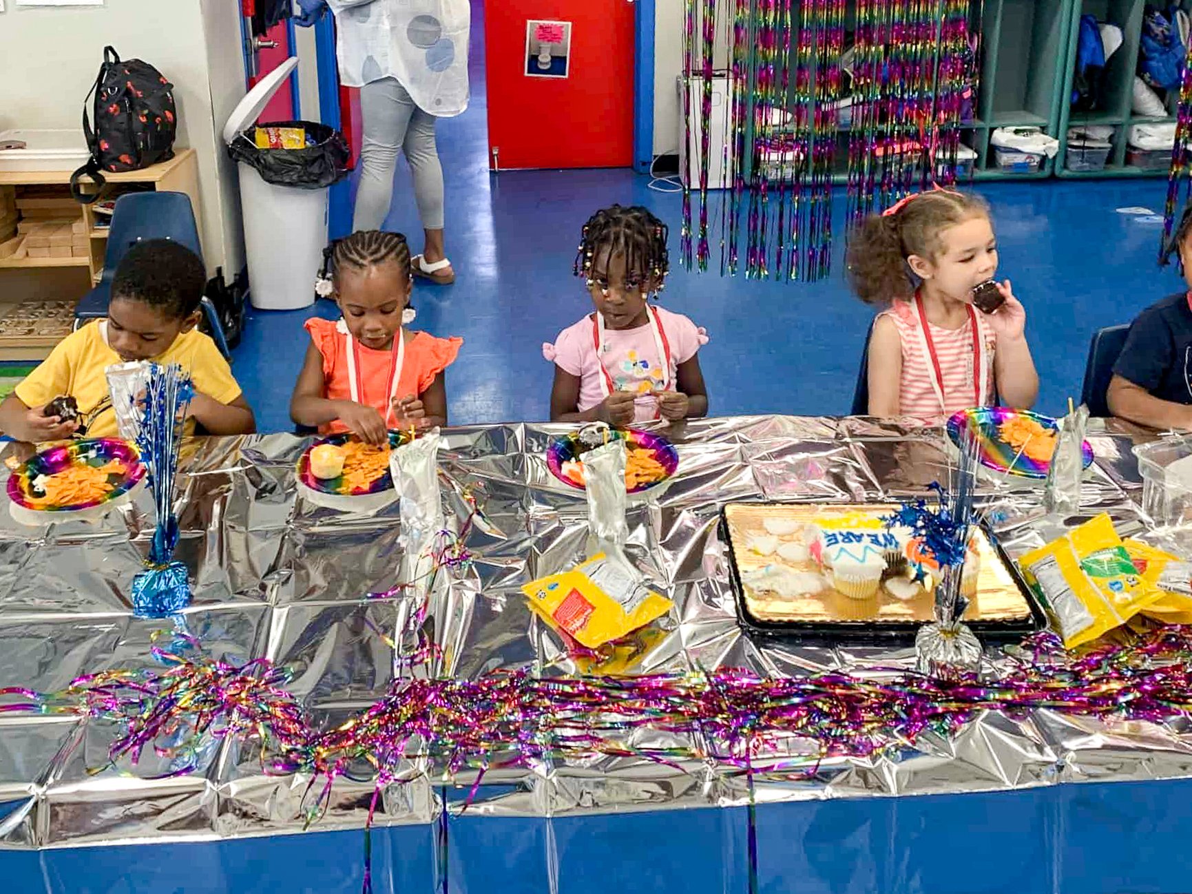 Children sitting at a party table with colorful decorations, cupcakes, and a cake, celebrating inside a room with blue flooring and storage shelves in the background at Tots Yearn to Learn.