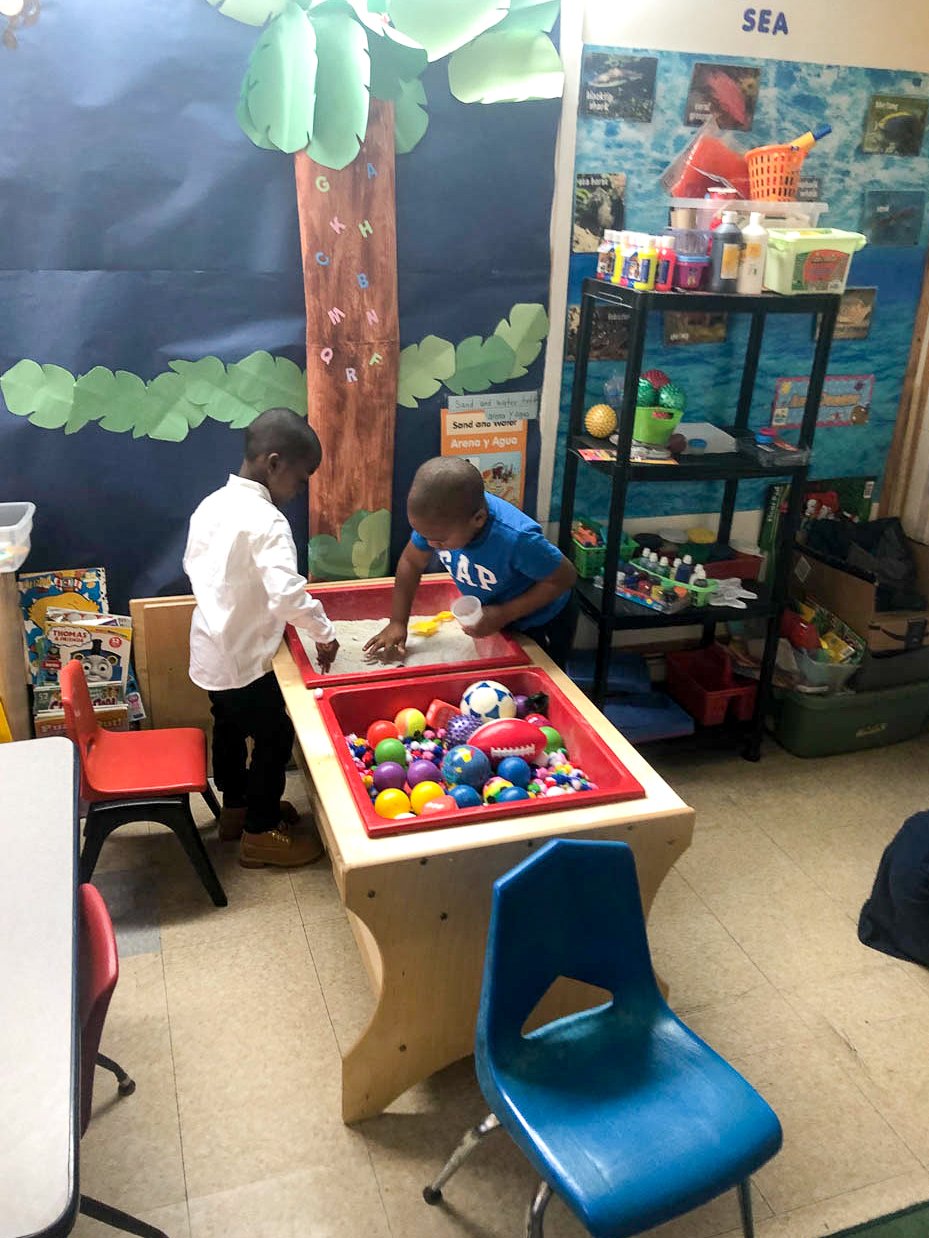 Two children playing at a sandbox table filled with colorful plastic balls and toys, in a Tots Yearn to Learn classroom decorated with a rainforest theme and educational posters.