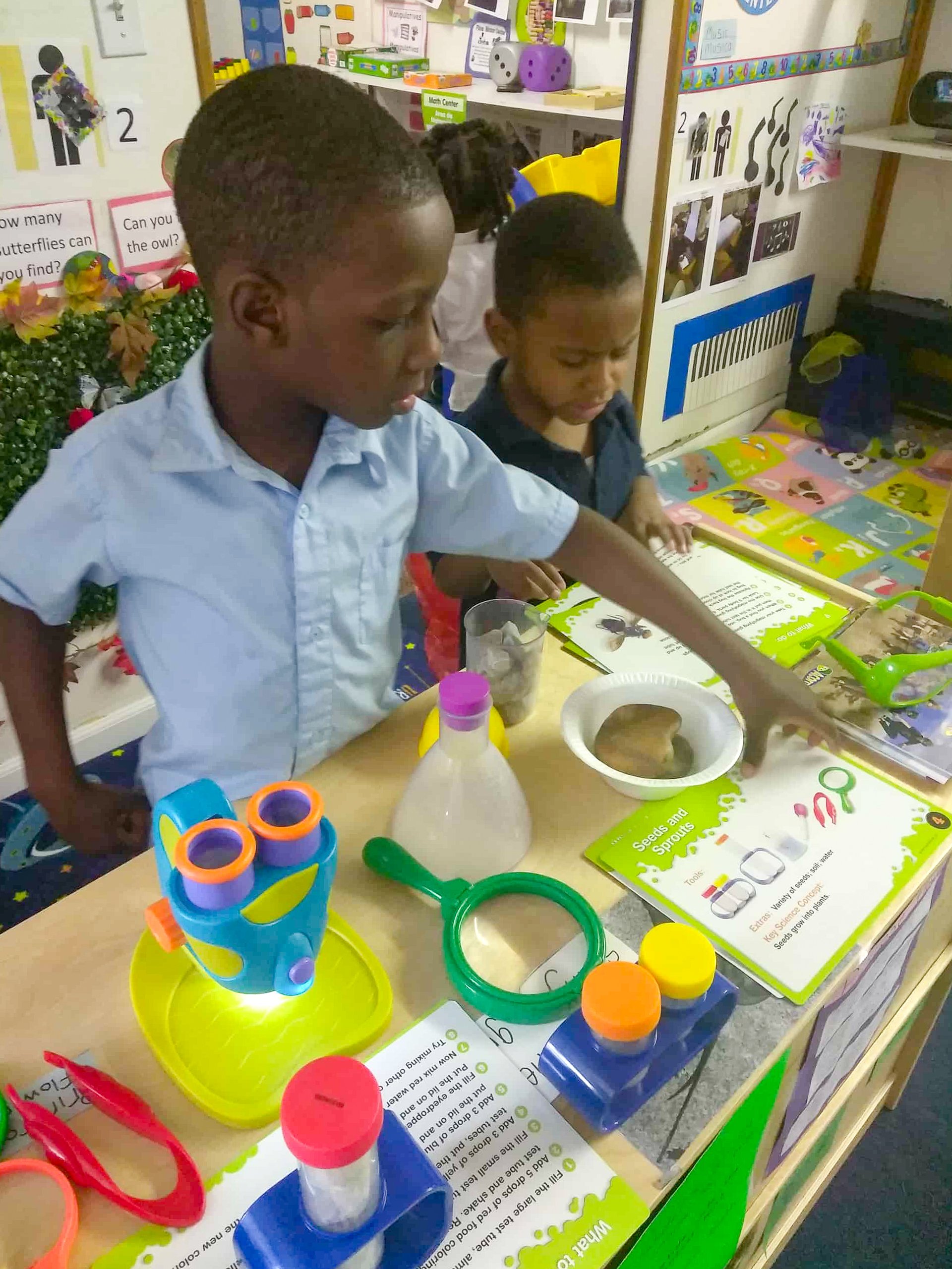 Two children are conducting a science experiment at a Tots Yearn to Learn classroom table with colorful science tools, books, and educational materials around them.