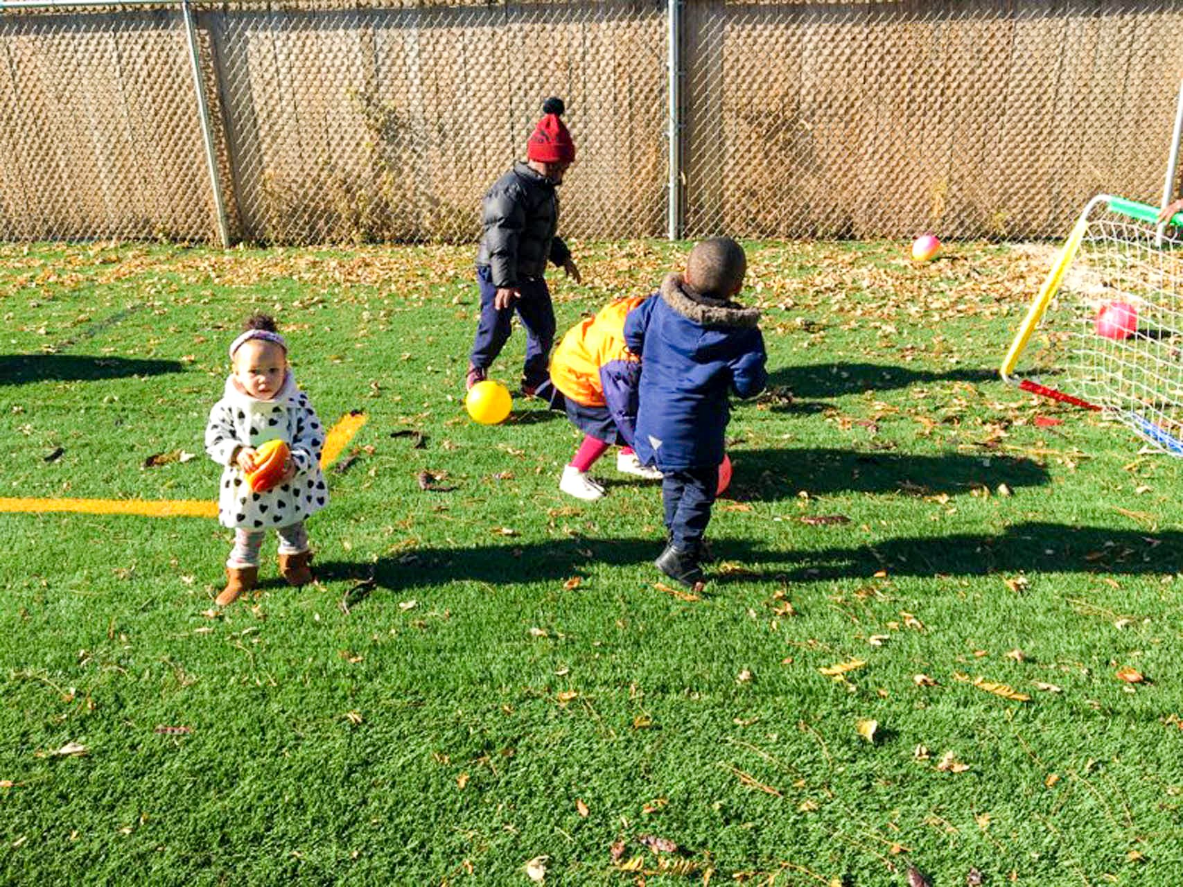 Several children playing soccer outdoors at Tots Yearn to Learn on a sunny day, with a fence and autumn leaves in the background.
