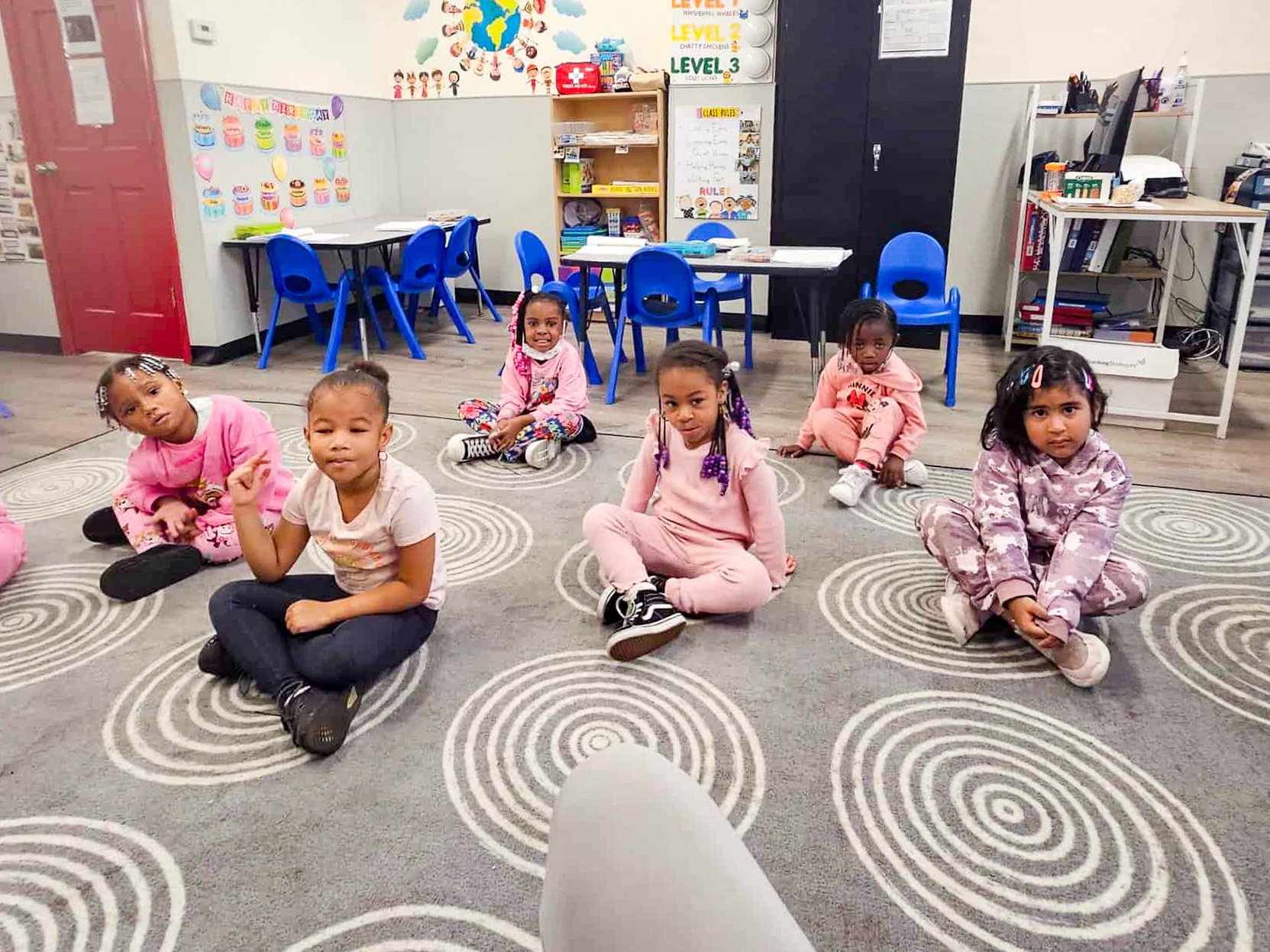 Six young girls sitting on a patterned carpet in a Tots Yearn to Learn classroom, some with braided hair and colorful clothing, with classroom furniture and educational posters in the background.