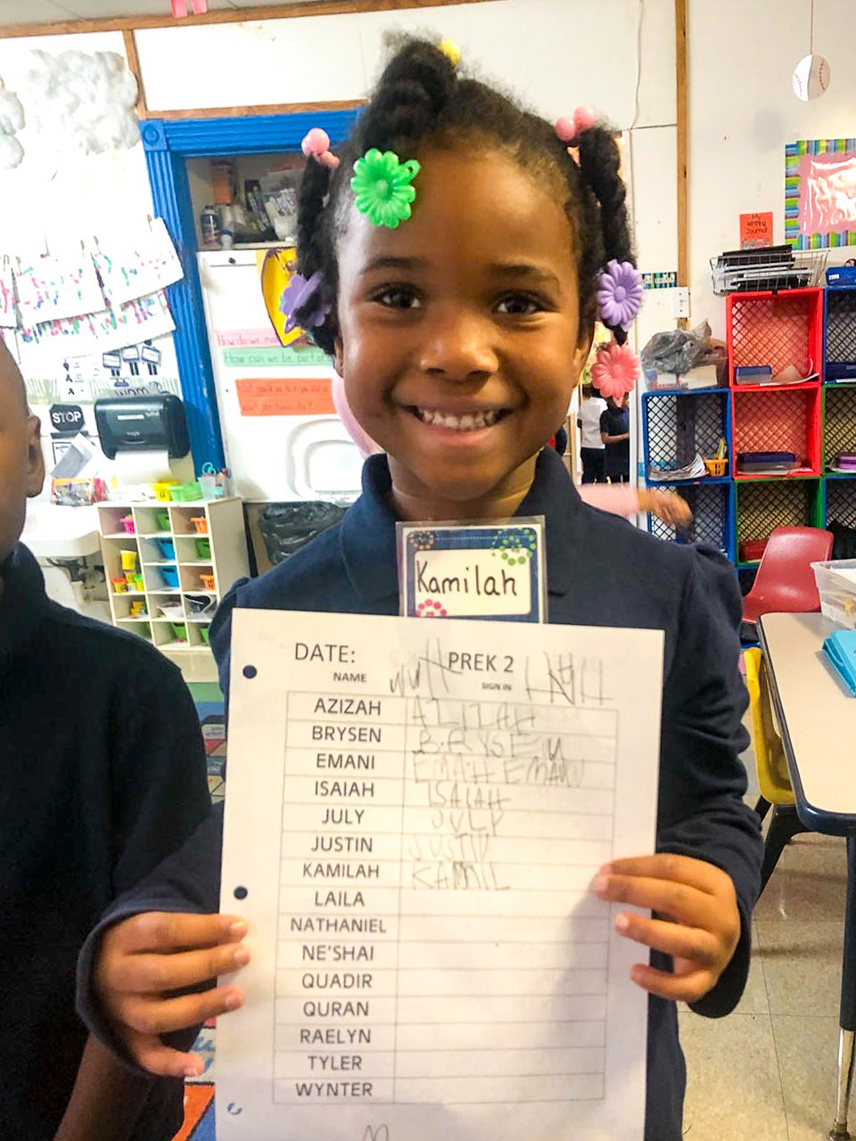 A young girl with braided hair decorated with colorful elastic bands and flowers, smiling and holding a classwork sheet in a classroom.