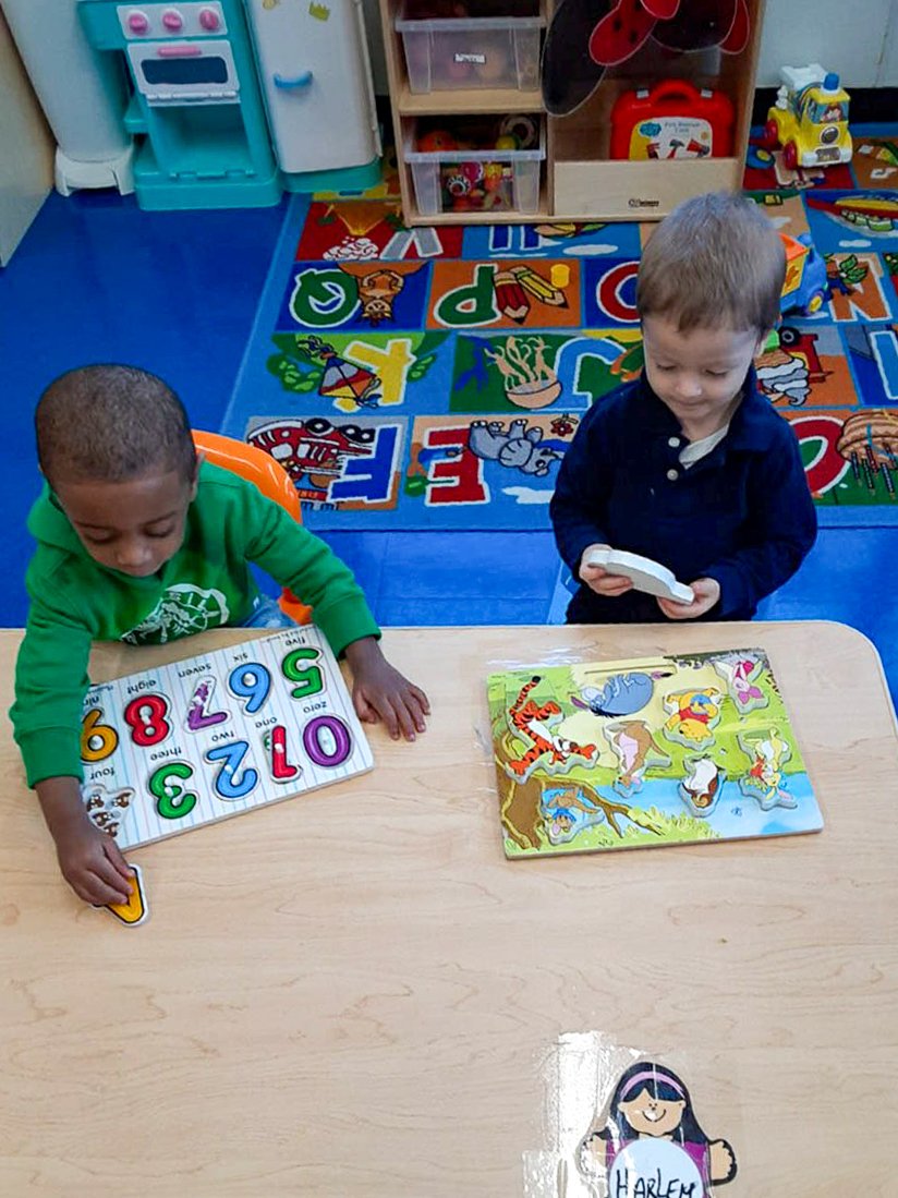 Two young boys sitting at a Tots Yearn to Learn classroom table playing with educational toys, surrounded by a colorful rug with alphabet and animal pictures in a classroom setting.