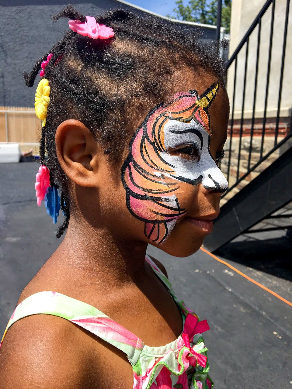 A young girl with pink, yellow, and blue hair clips, wearing a colorful, floral dress, has her face painted as a unicorn with pink, orange, white, and black details, and is smiling with closed eyes outdoors.