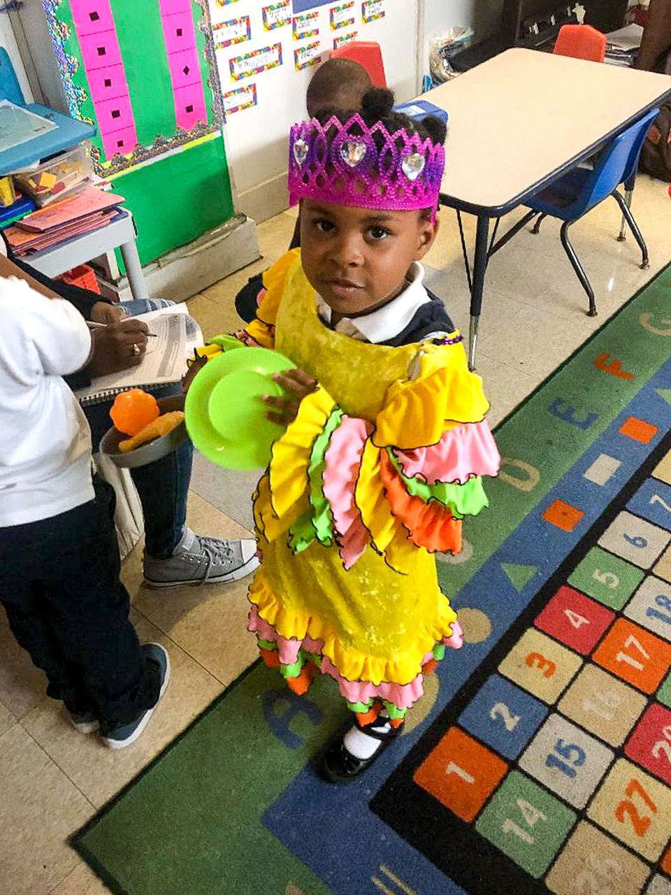 A young girl dressed in a colorful, ruffled dress with a pink crown decorated with heart shapes, standing on a classroom rug with numbers and letters, looking at the camera.