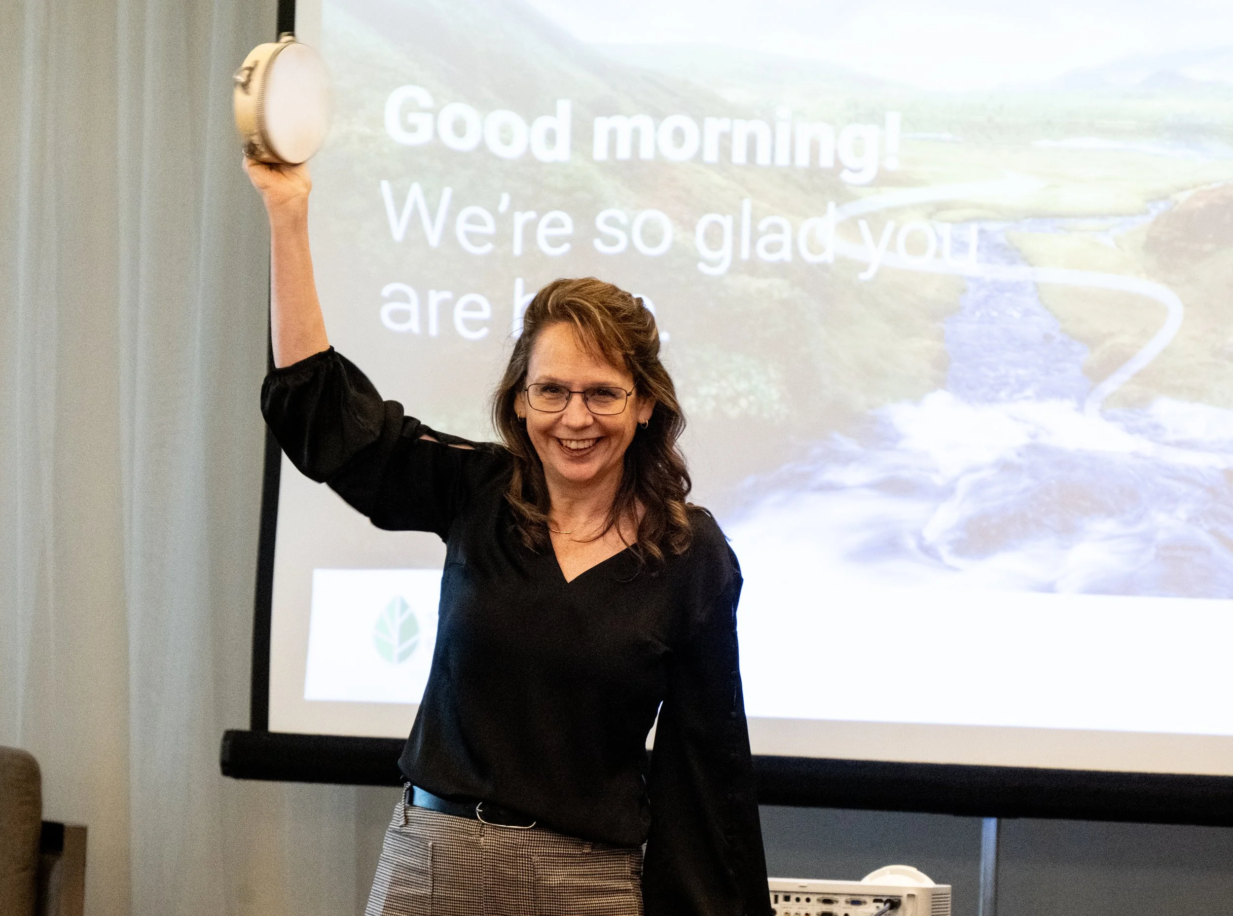 Nancy Wise smiling and holding a tambourine  in front of a projection screen with outdoor scenery and text that says 'Good morning! We're so glad you are here'.