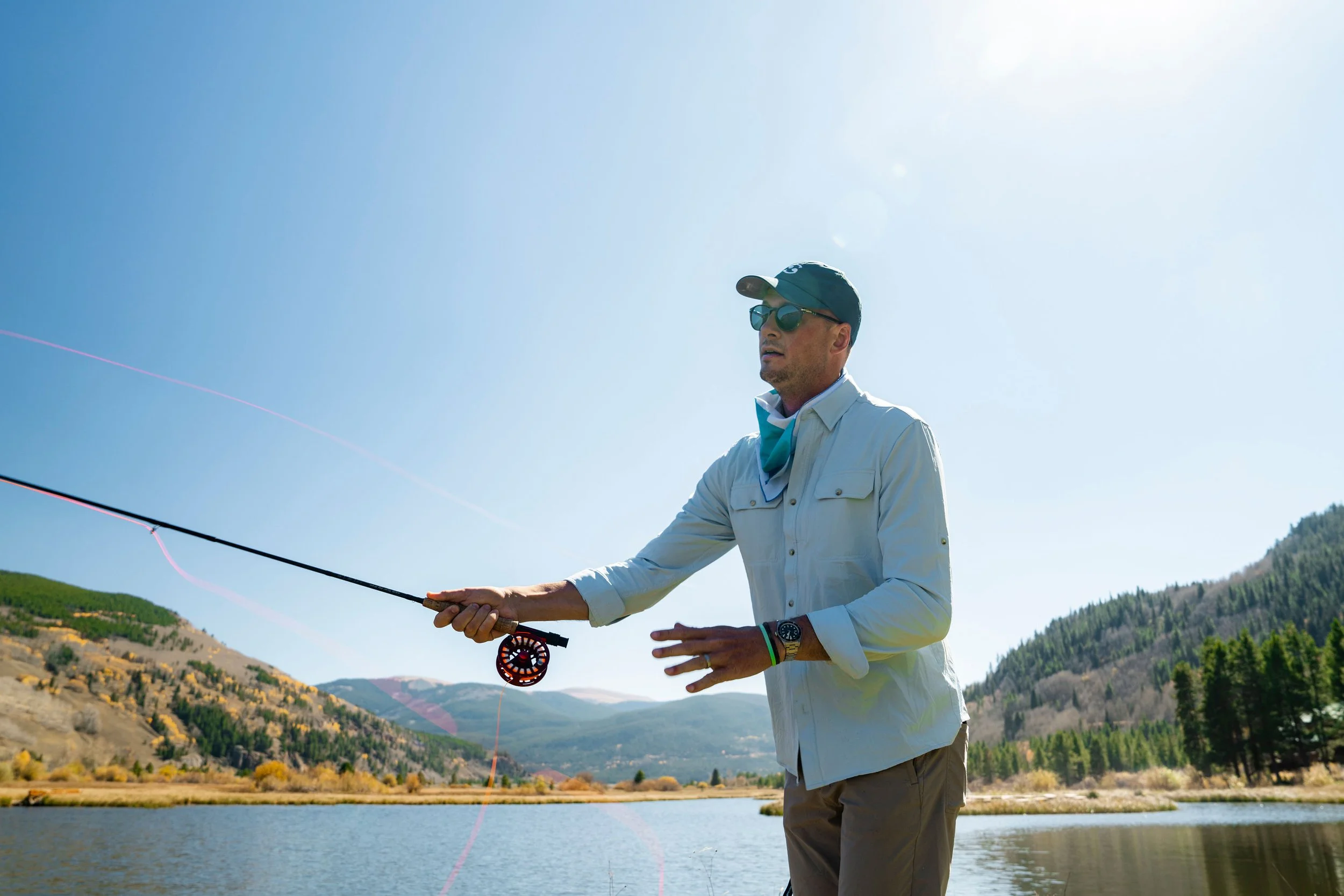 Man fishing in a lake surrounded by mountains and trees on a sunny day.