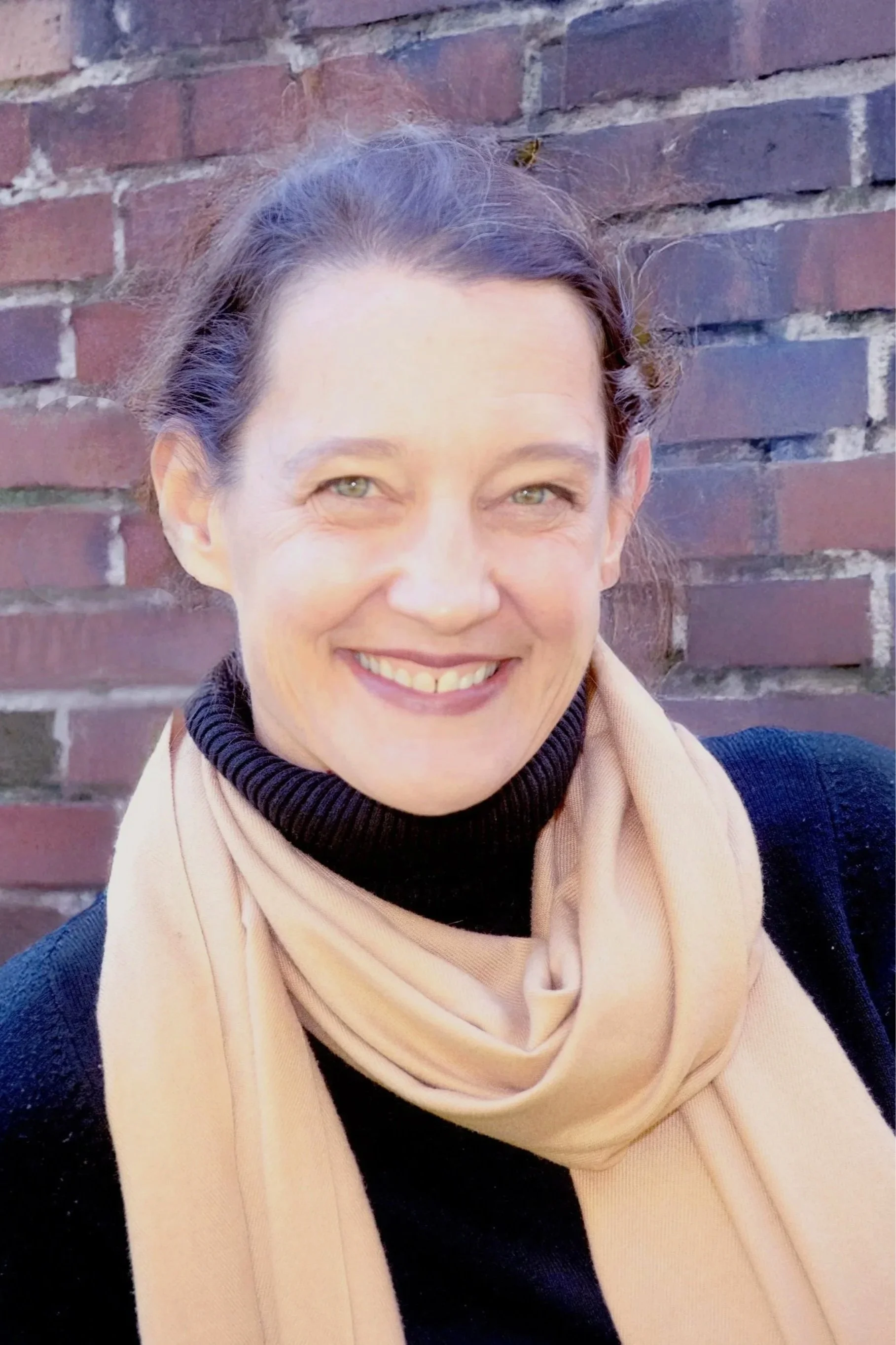 Smiling woman outdoors in front of a brick wall, wearing a black turtleneck and a beige scarf.