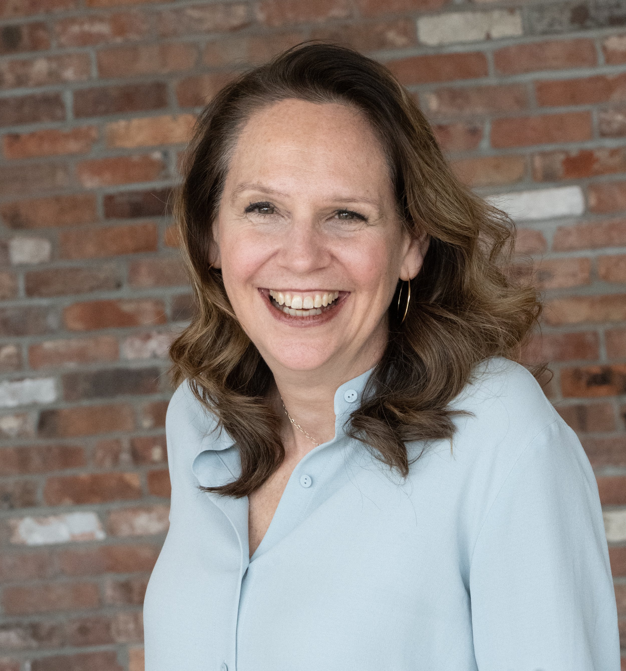 A woman with shoulder-length brown hair, wearing a light blue blouse, smiling, standing in front of a brick wall.